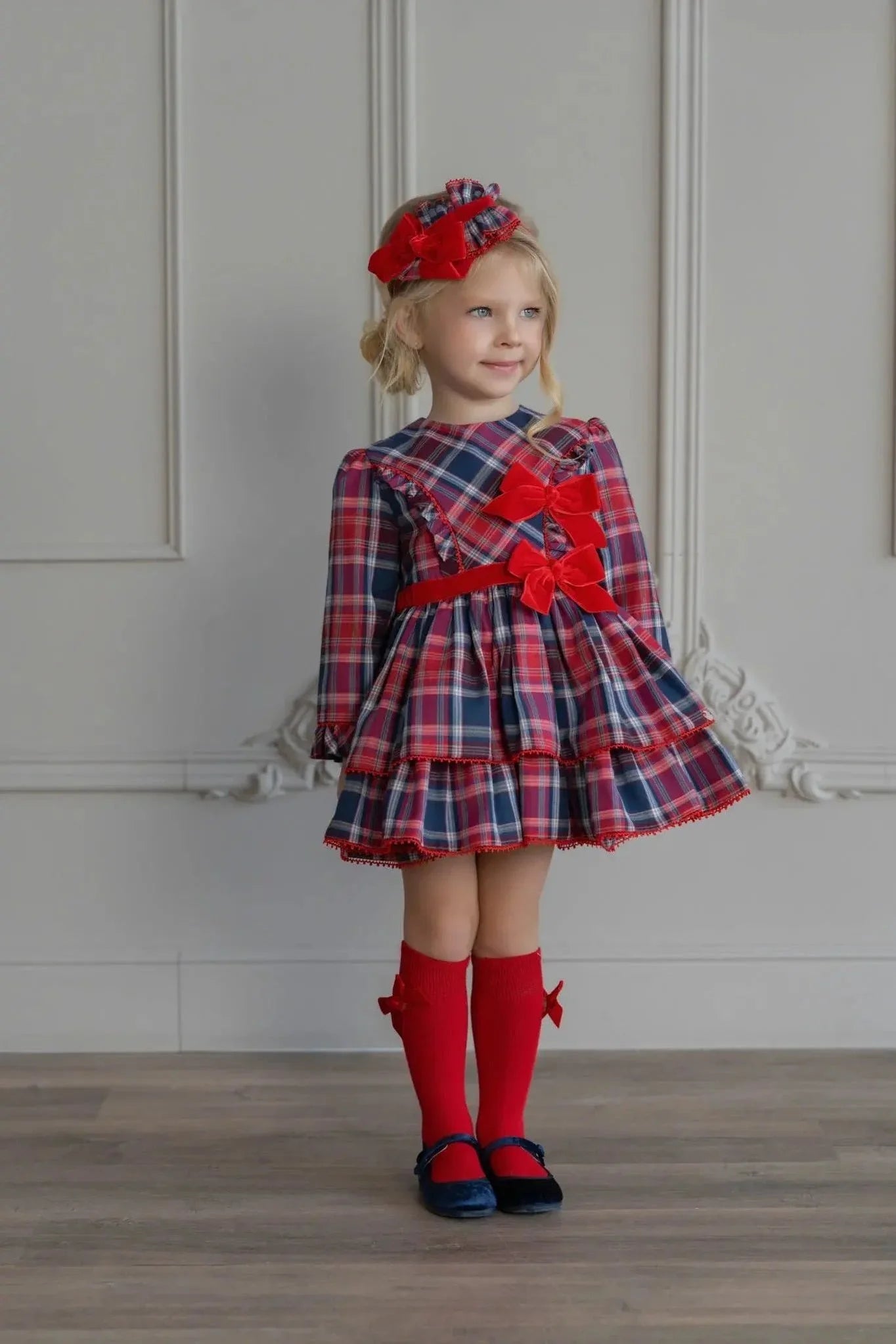 Little girl in a red and navy plaid dress with red bows, matching headband, red knee socks, and navy shoes indoors