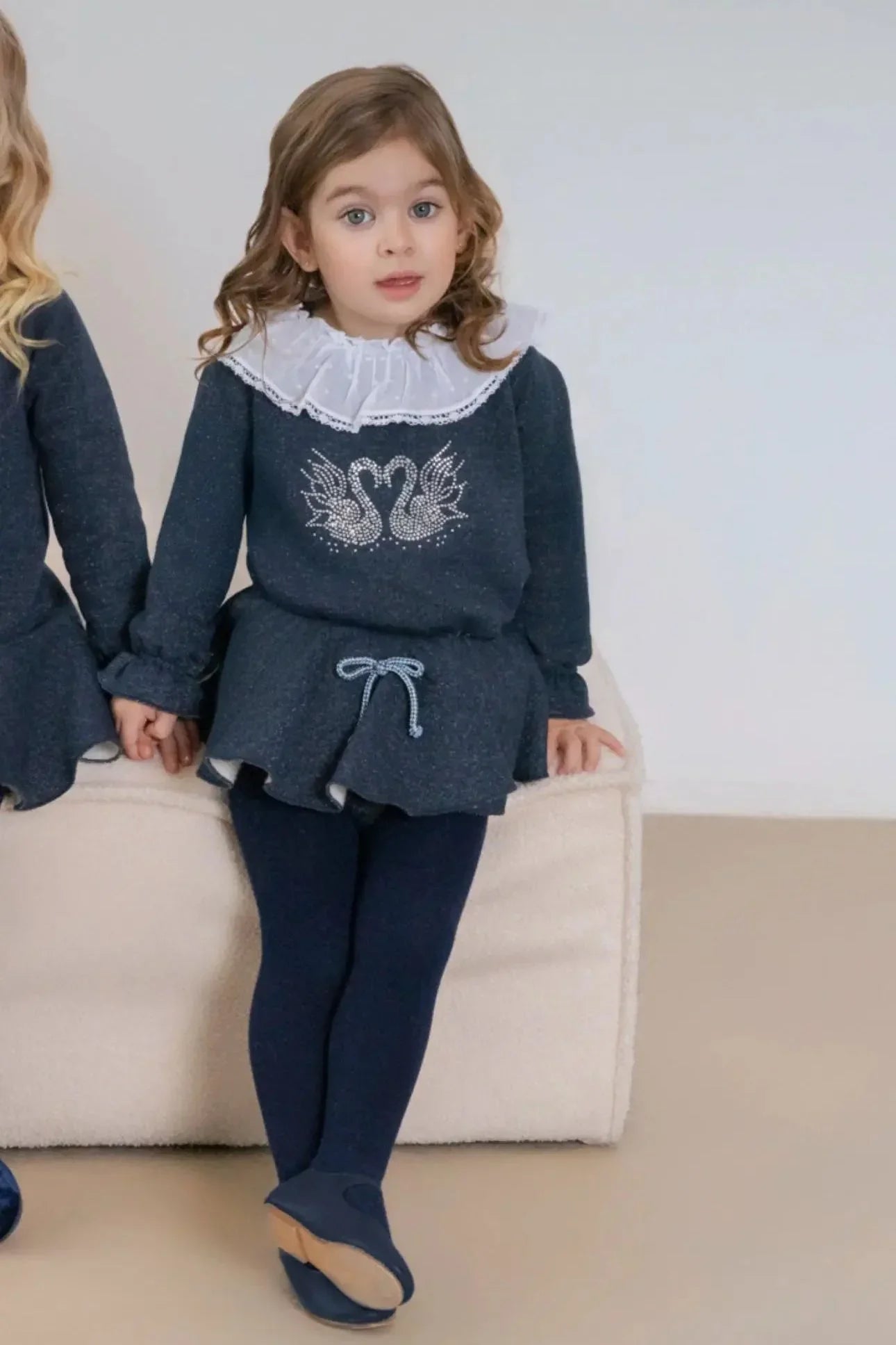 Young girl in navy swan print top with lace collar, matching skirt and tights, sitting on beige couch