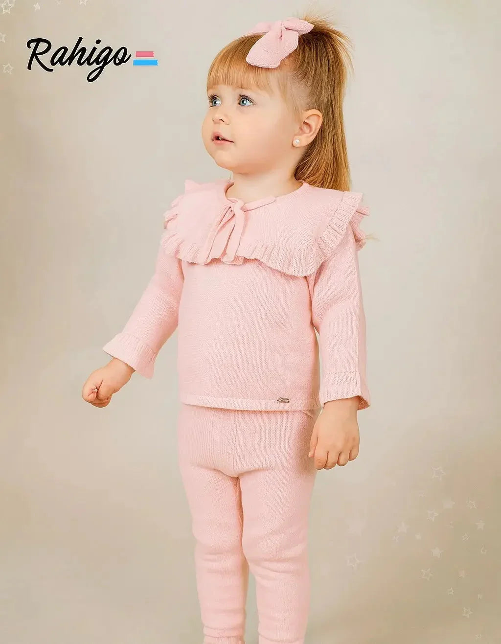 Toddler girl wearing a pink knitted tracksuit with ruffled collar and bow headband on beige background