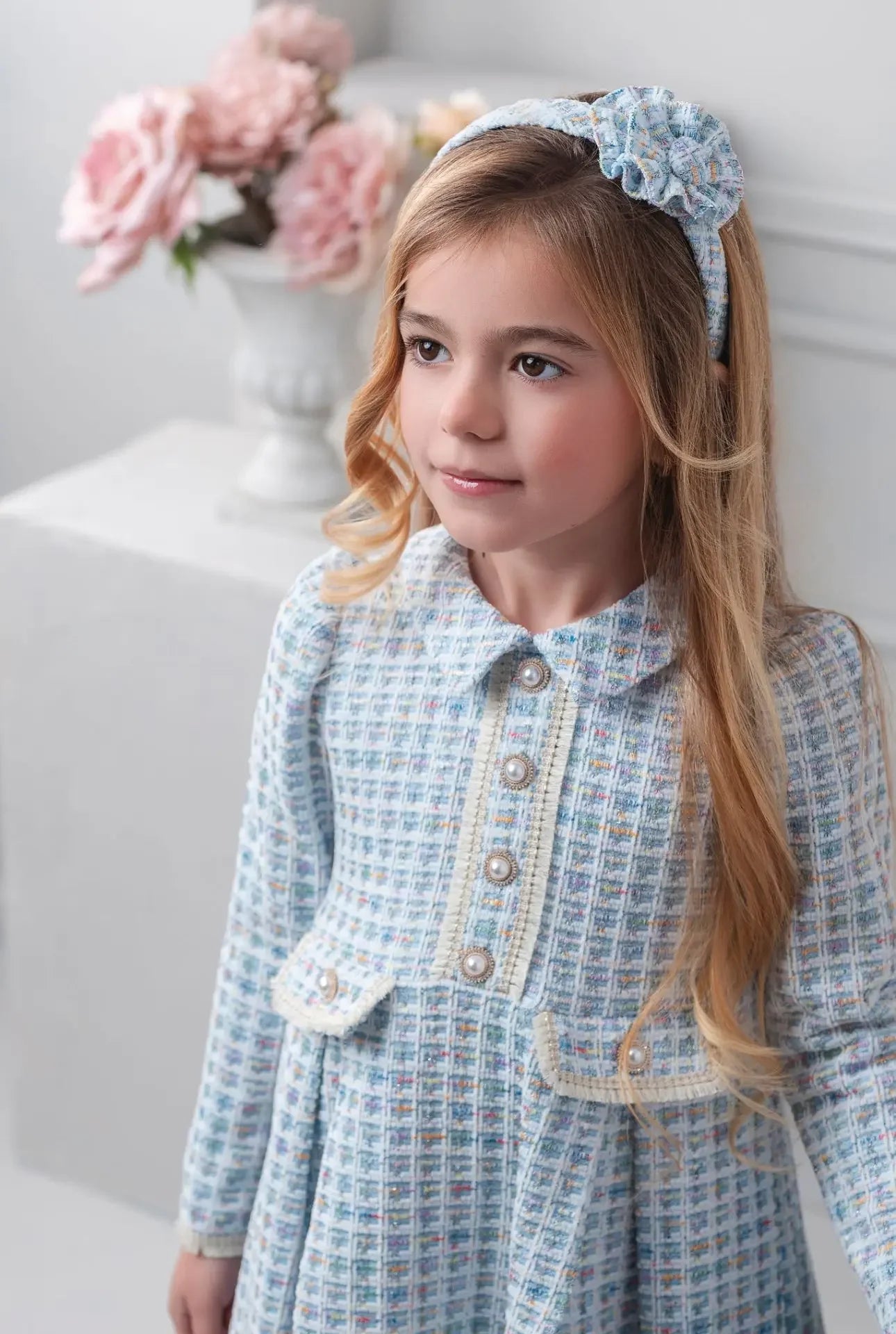 Young girl in blue tweed dress with pearl buttons and matching floral headband, soft pink flowers background