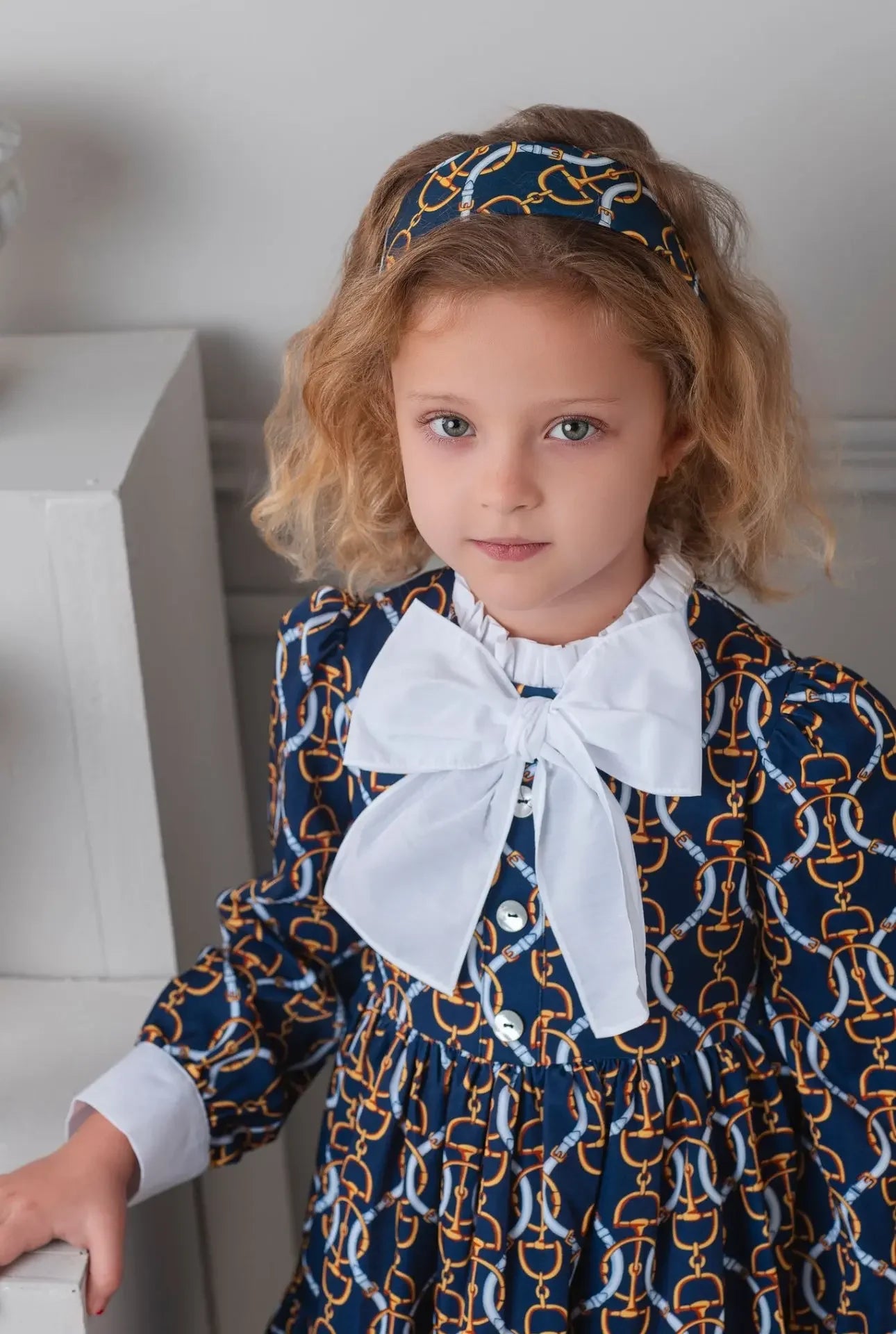 Young girl wearing navy dress with gold chain print and large white bow, posing indoors