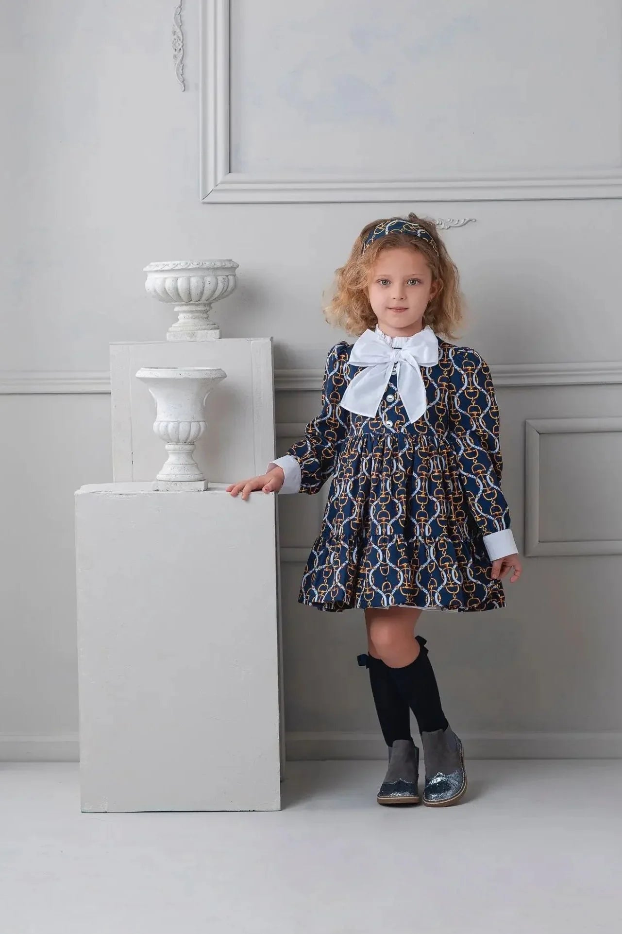 Young girl in navy blue dress with gold chain print, white bow, and knee-high socks standing by white pedestals