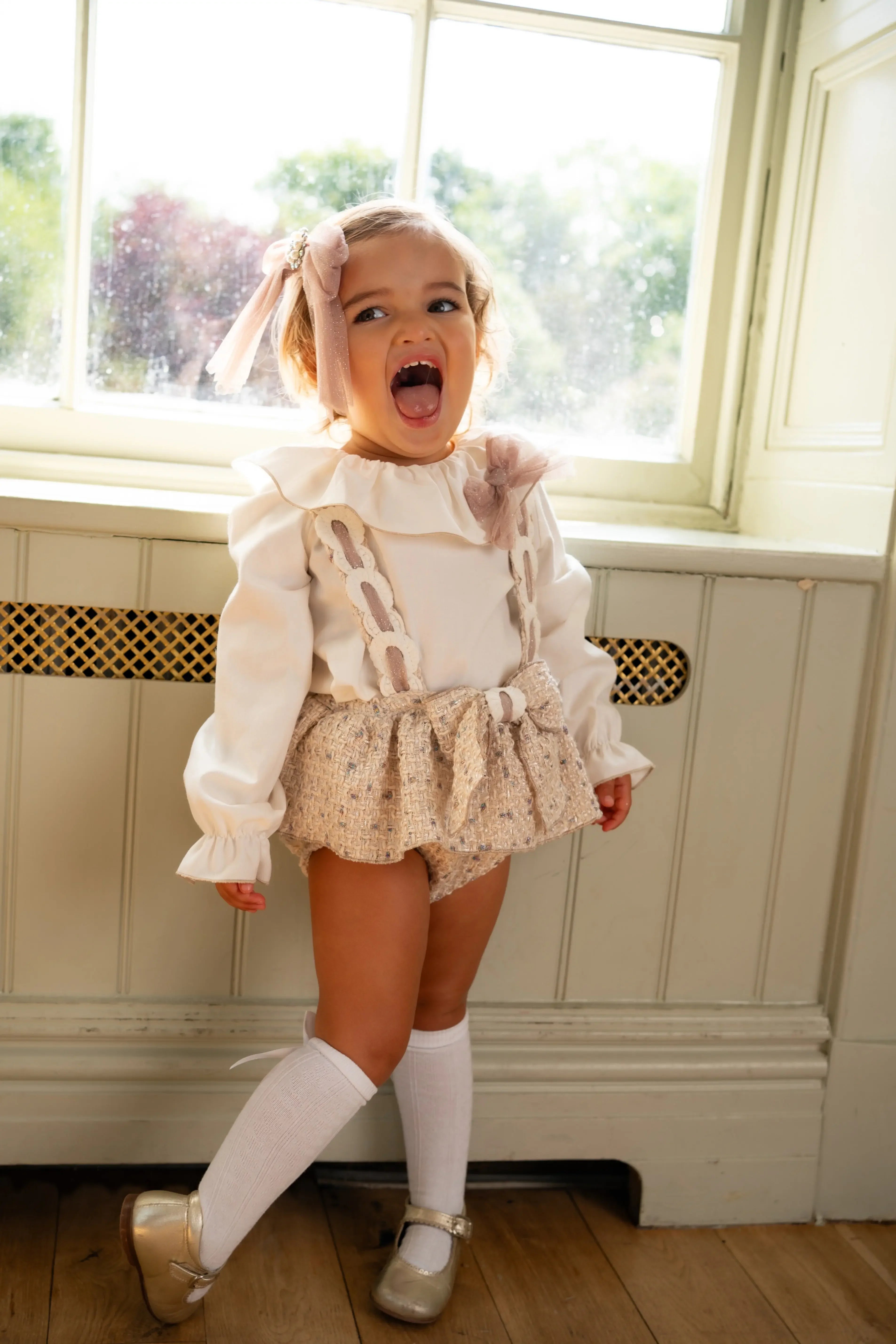 Toddler girl in beige skirt, white blouse, knee-high socks, and gold shoes by window