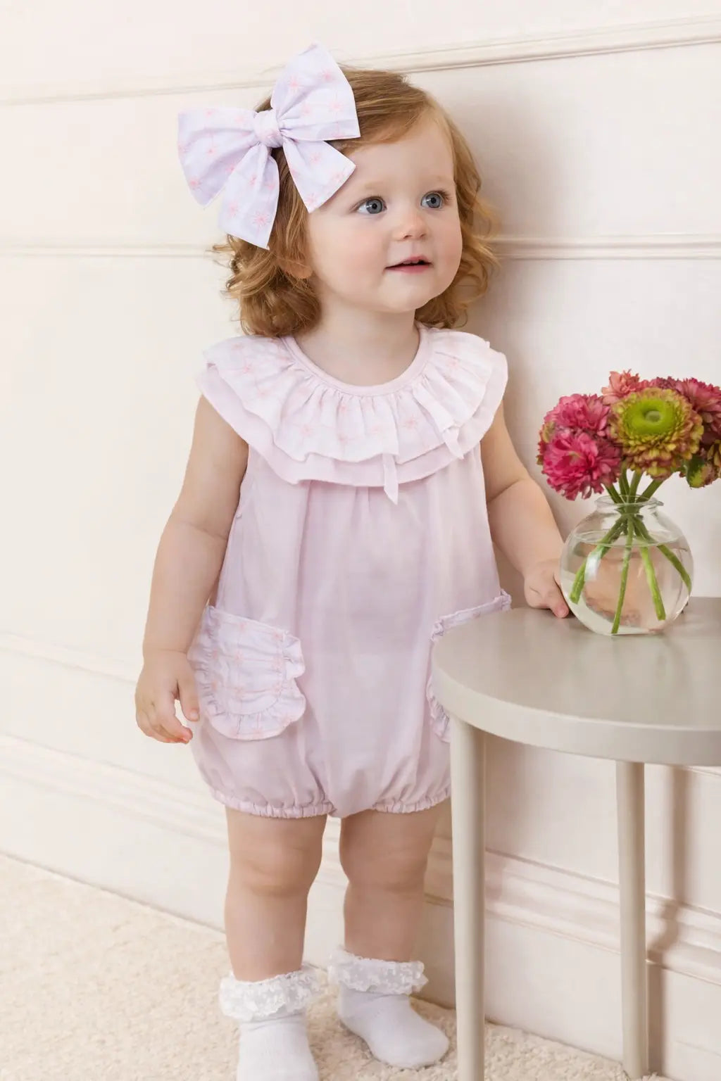 Young child in a pink outfit with a large bow, standing next to a small table with a vase of flowers.
