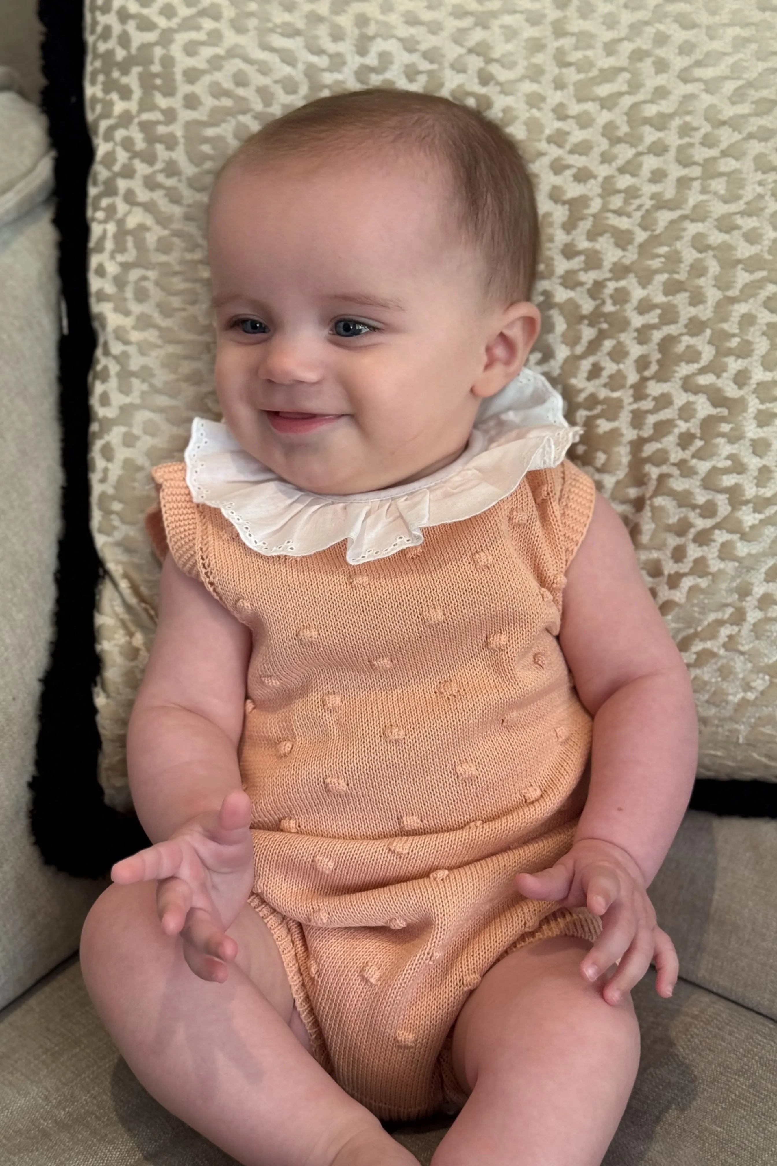 Baby girl smiling wearing peach knitted romper with white frill collar sitting on patterned cushion
