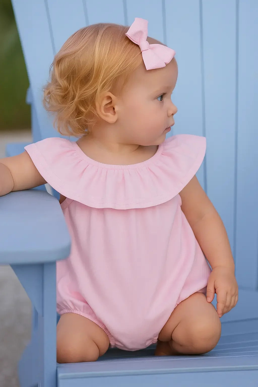 Baby girl in pink linen summer romper with ruffle collar and pink bow sitting on light blue chair