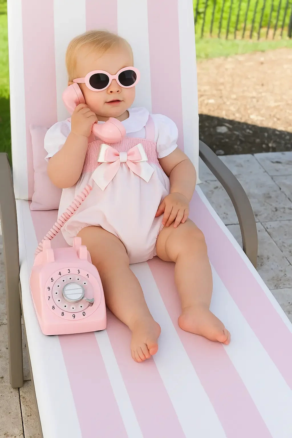 Baby girl wearing pink sunglasses and white pink summer romper sitting on striped lounge chair holding pink rotary phone