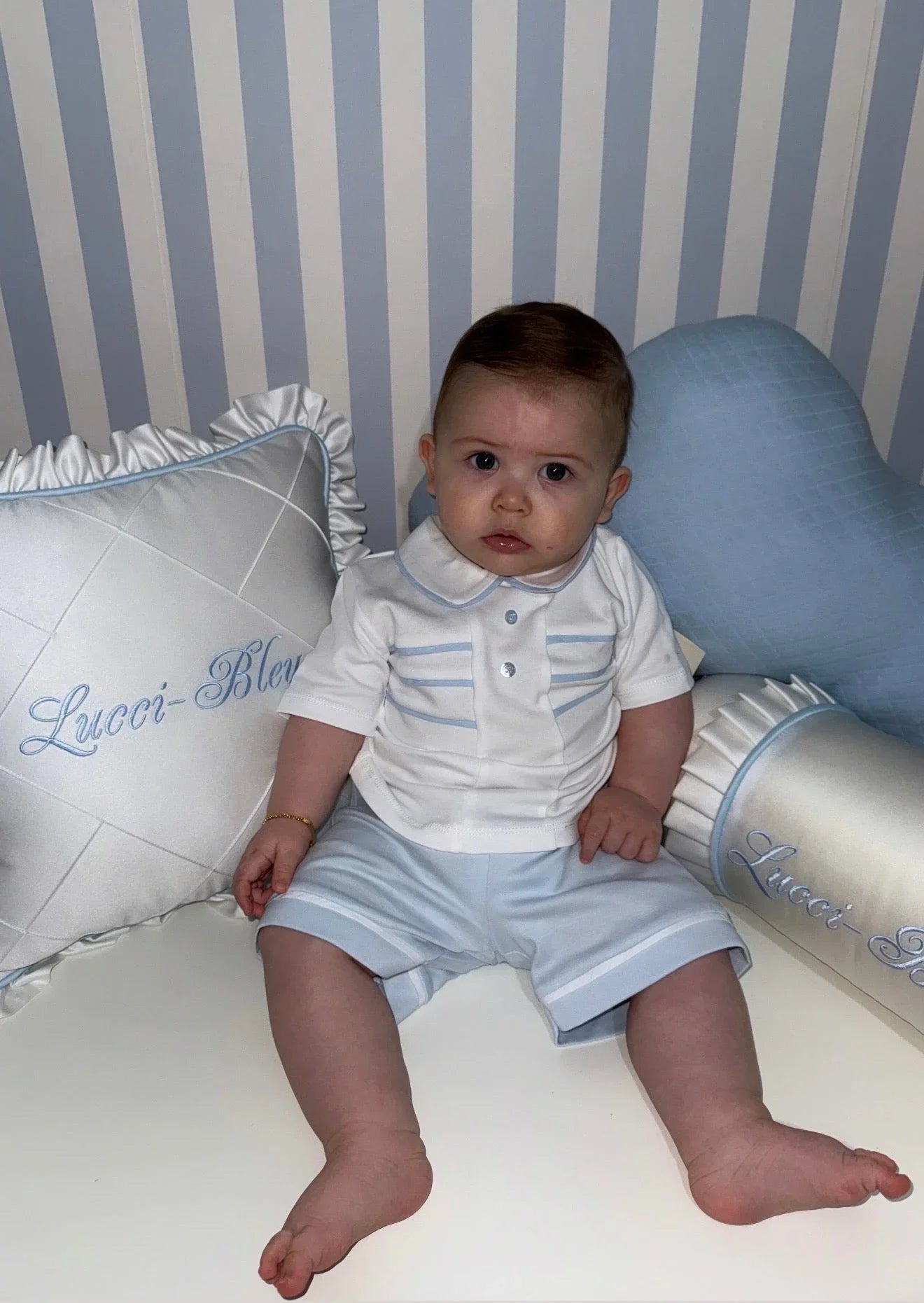Baby boy in white and light blue outfit sitting between two pillows embroidered with Lucçi-Bleu on striped background
