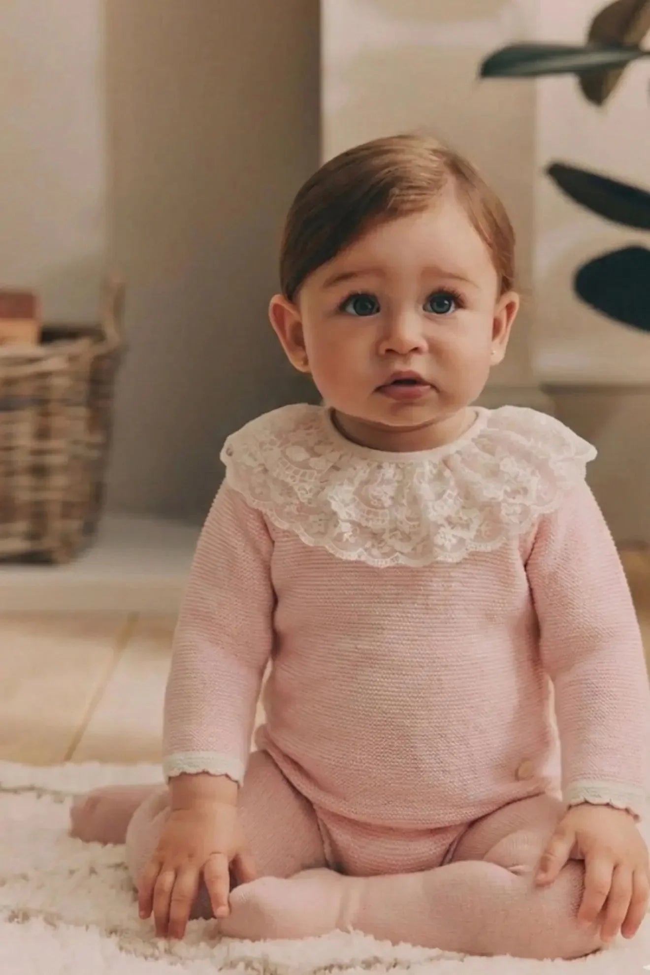 Baby girl with blue eyes sitting on a soft rug wearing a pink knit outfit with white lace collar