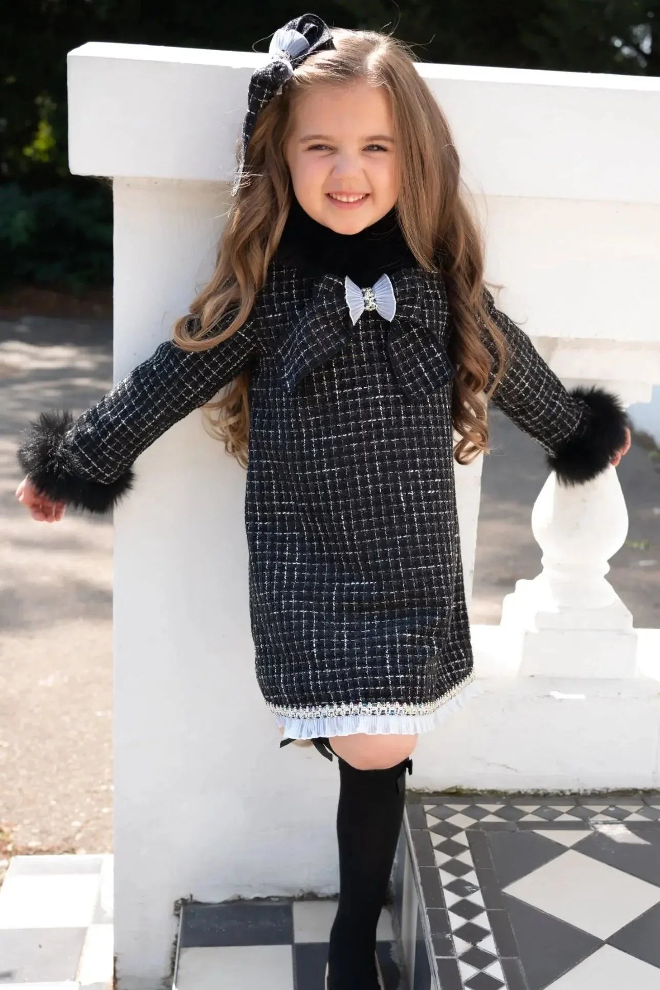 Smiling young girl in black and white checkered dress with fur cuffs and bow, standing outdoors