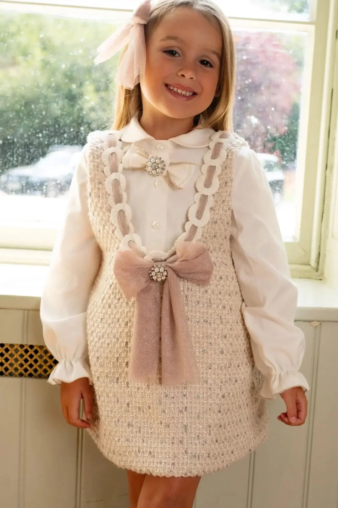 Smiling girl in beige tweed dress with pearl bows and white blouse by a window