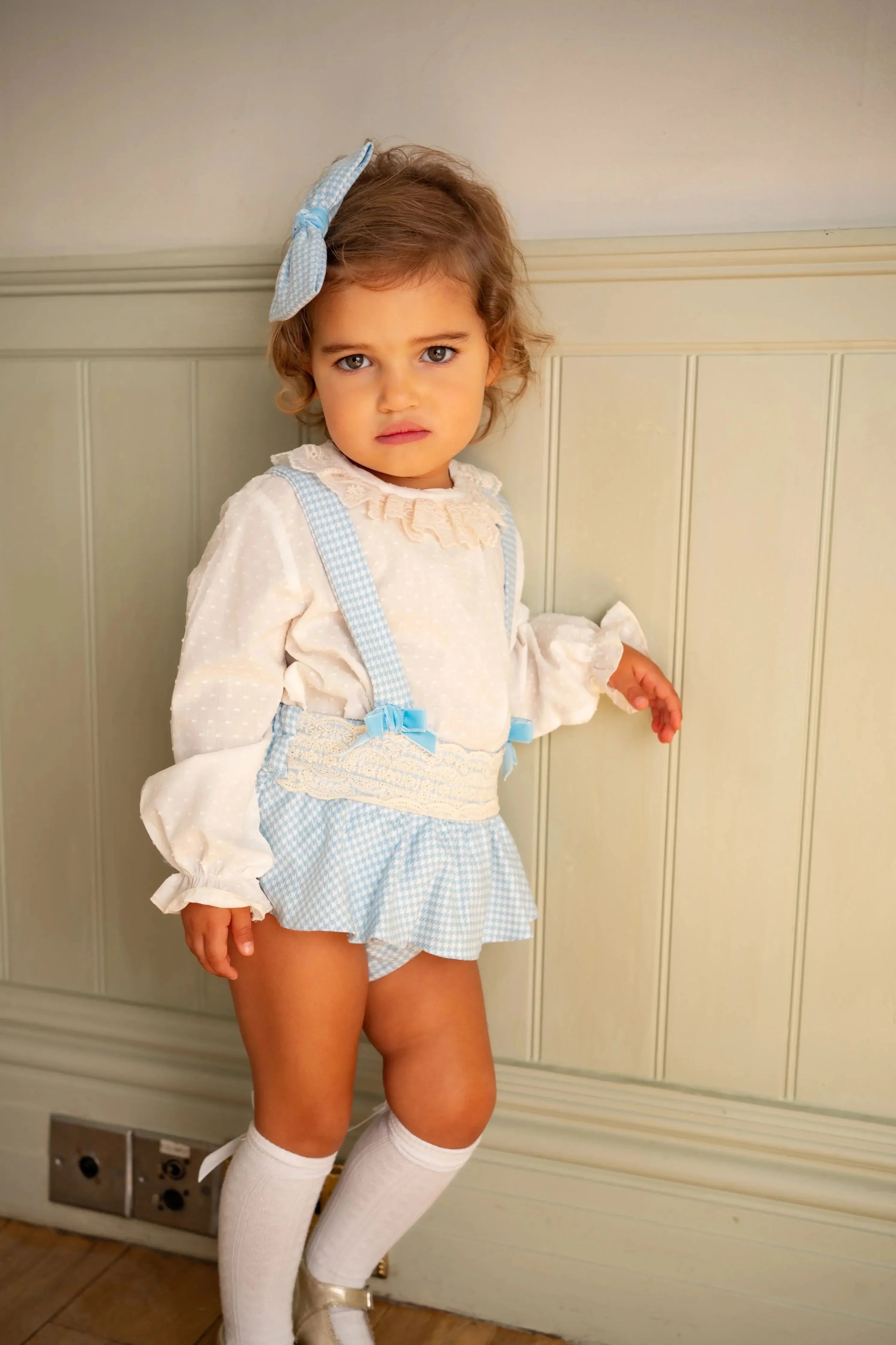 Toddler girl in blue gingham suspender skirt and white blouse with lace, standing indoors