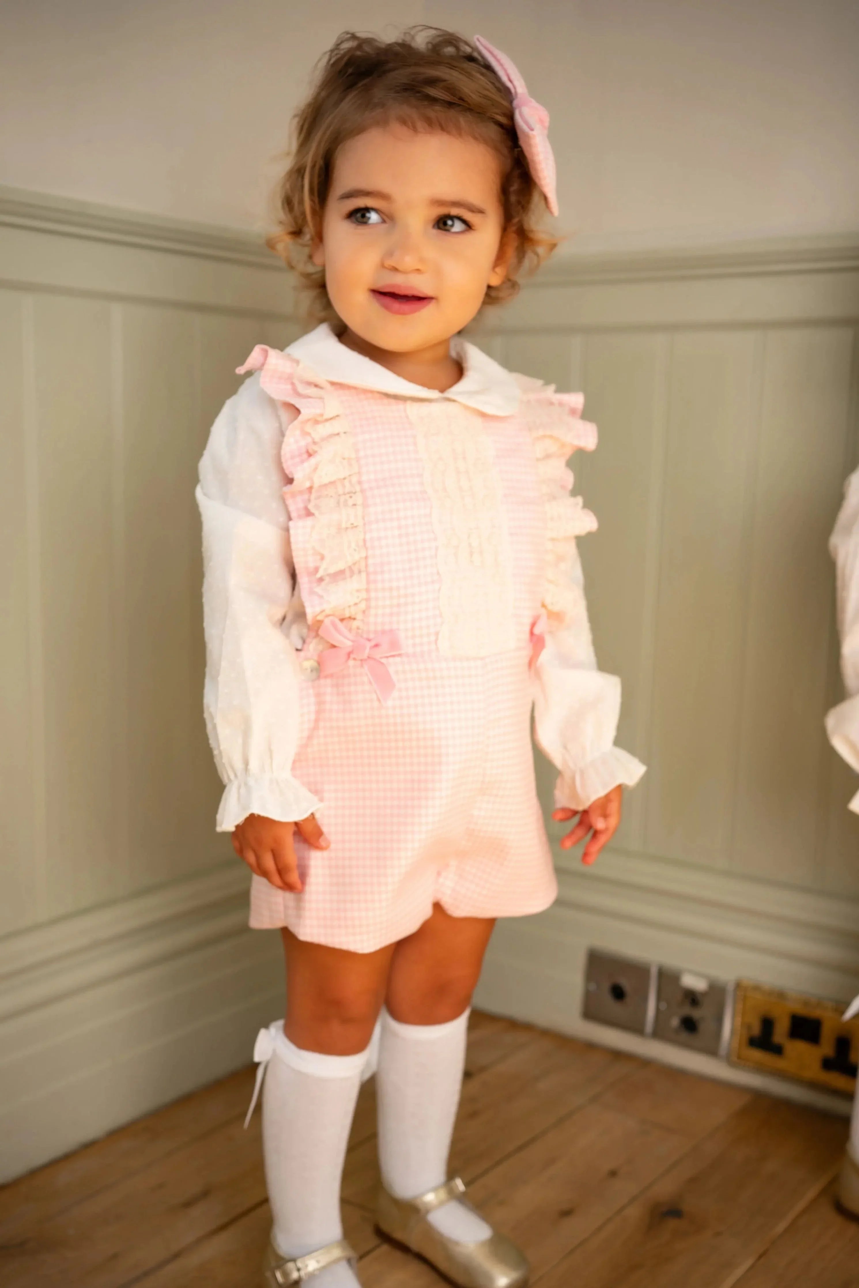 Toddler girl wearing pink gingham pinafore with ruffles, white blouse, knee-high socks, and gold shoes indoors