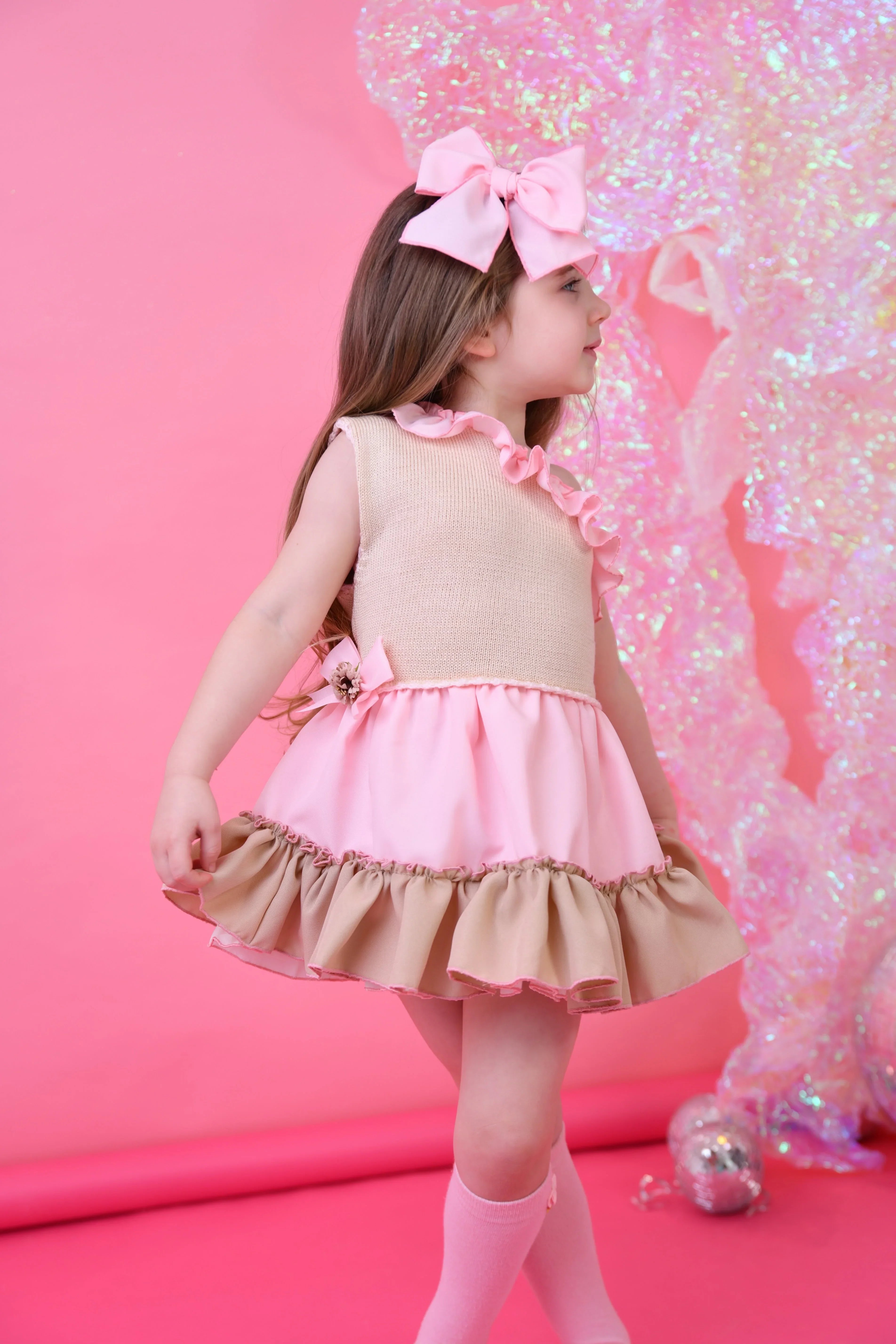 Little girl in pink and beige ruffled dress with large pink bow headband on pink background