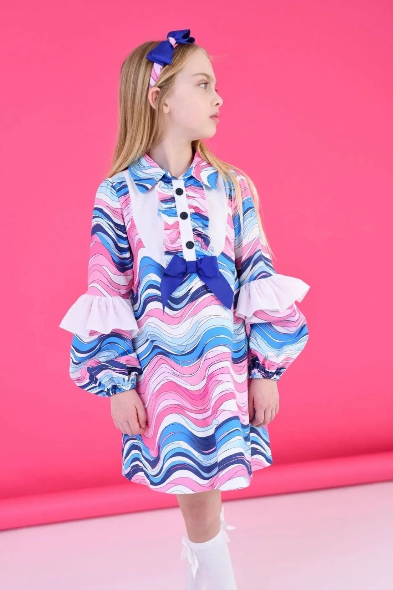 Girl in colorful wavy print dress with ruffled sleeves and blue bow against pink background