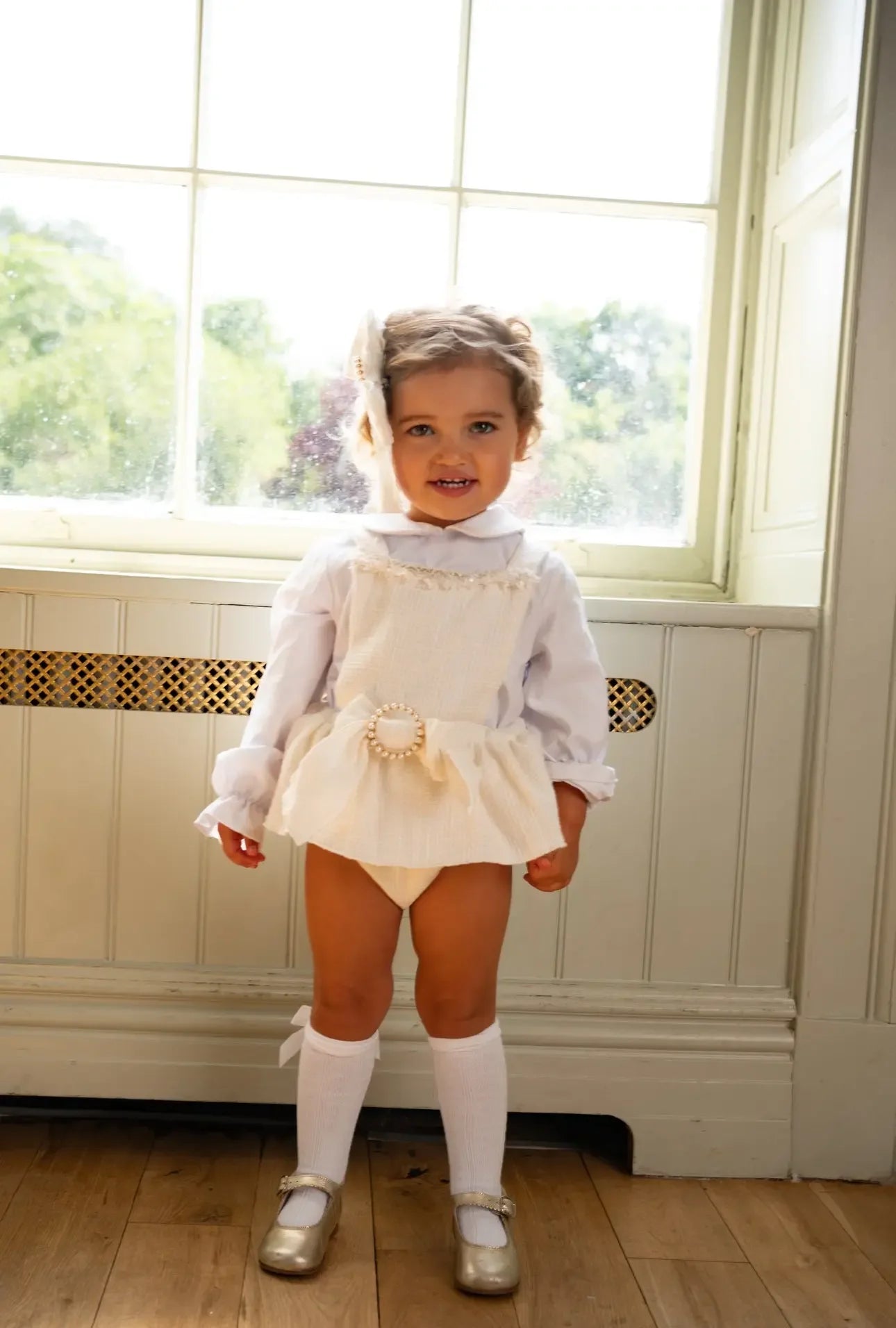 Toddler girl in white vintage outfit with pearl belt, knee-high socks, and gold shoes standing indoors