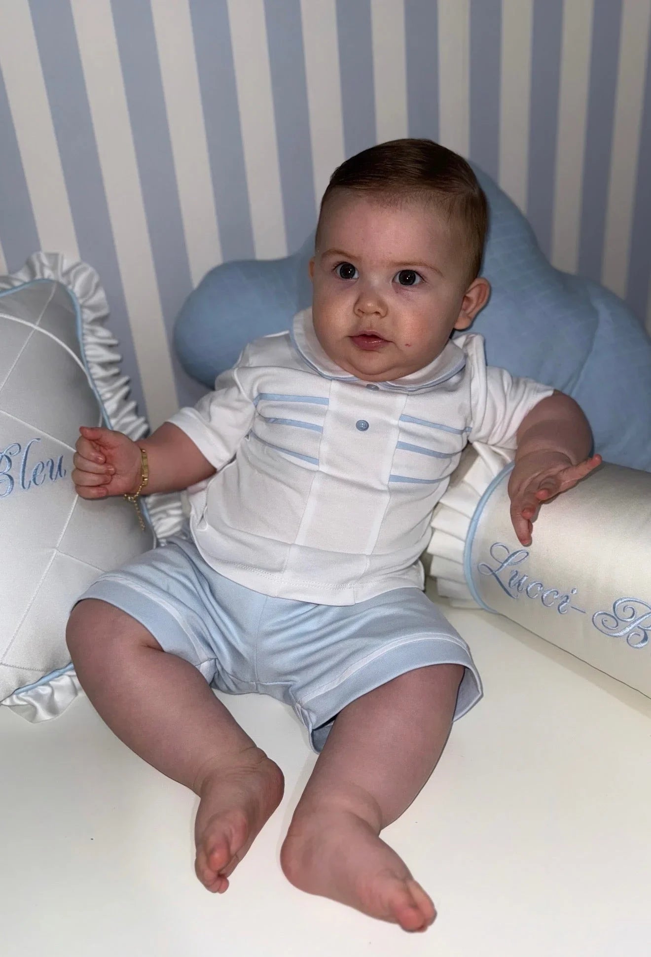 Baby boy in white and light blue outfit sitting on a white surface with blue striped background and decorative pillows