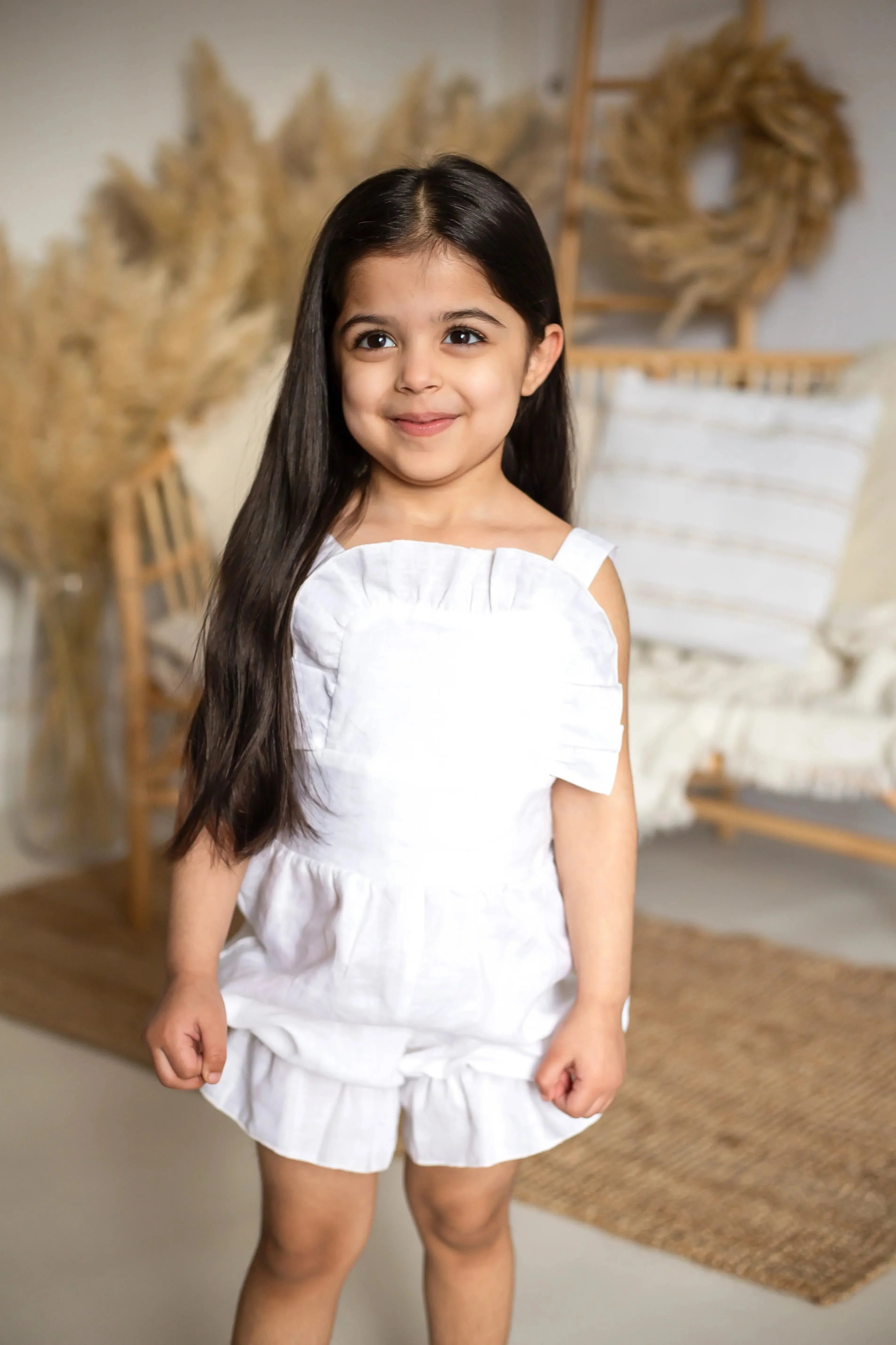 Smiling young girl with long dark hair wearing an ivory linen frilly jumpsuit in a cozy room