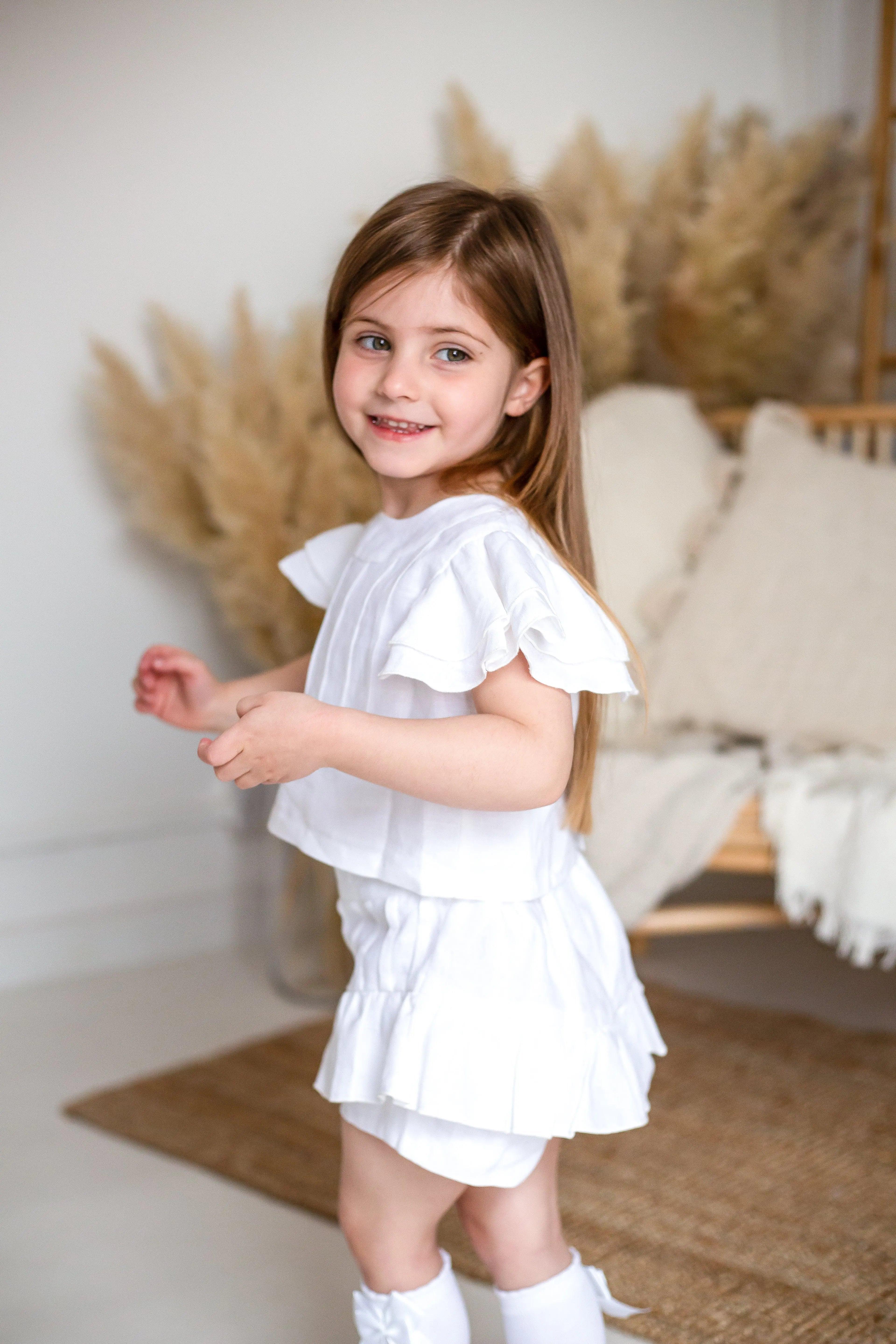 Smiling young girl in ivory linen pleated short set with ruffled sleeves in cozy neutral room