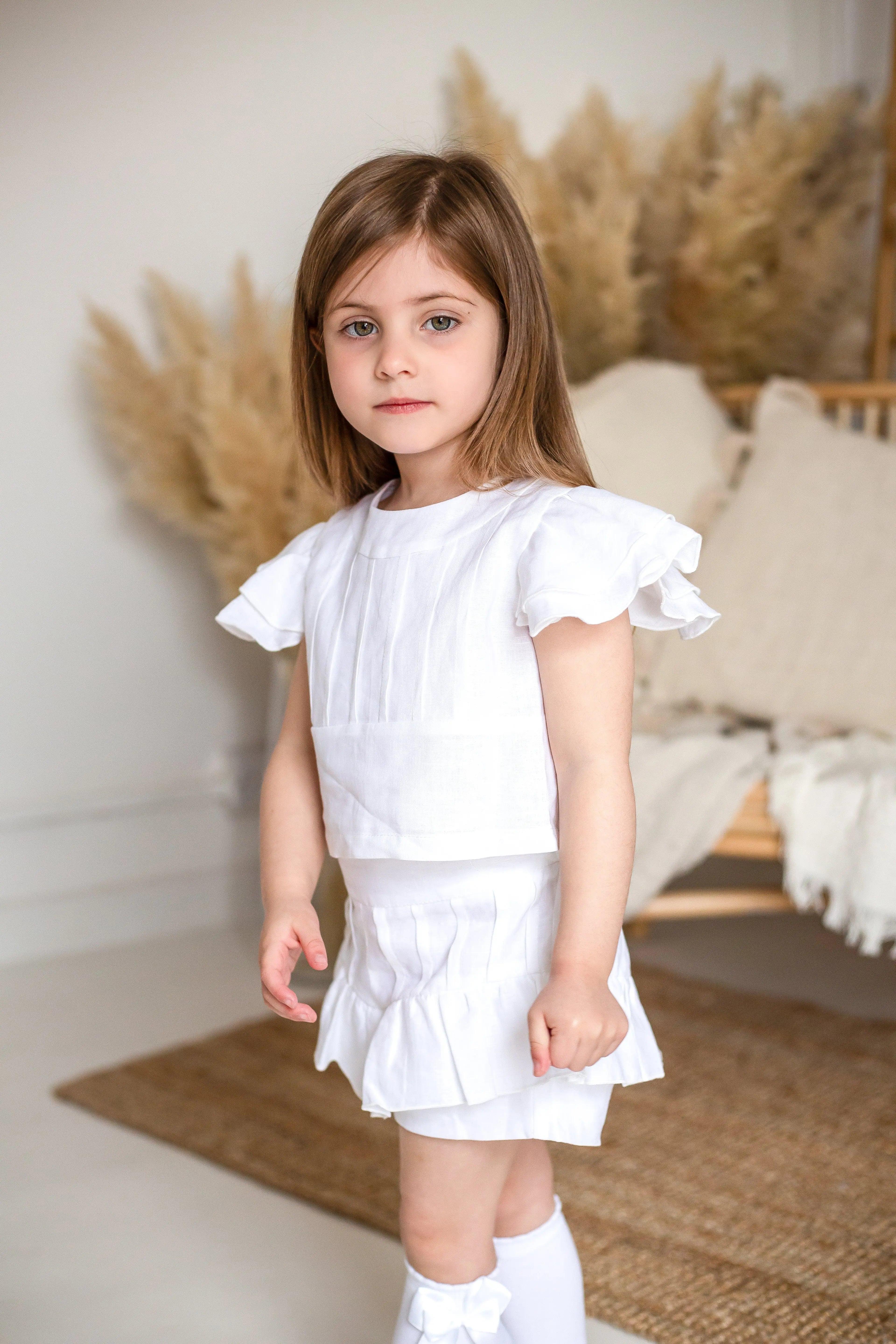 Young girl in white ruffled dress and knee-high socks standing indoors with neutral decor background