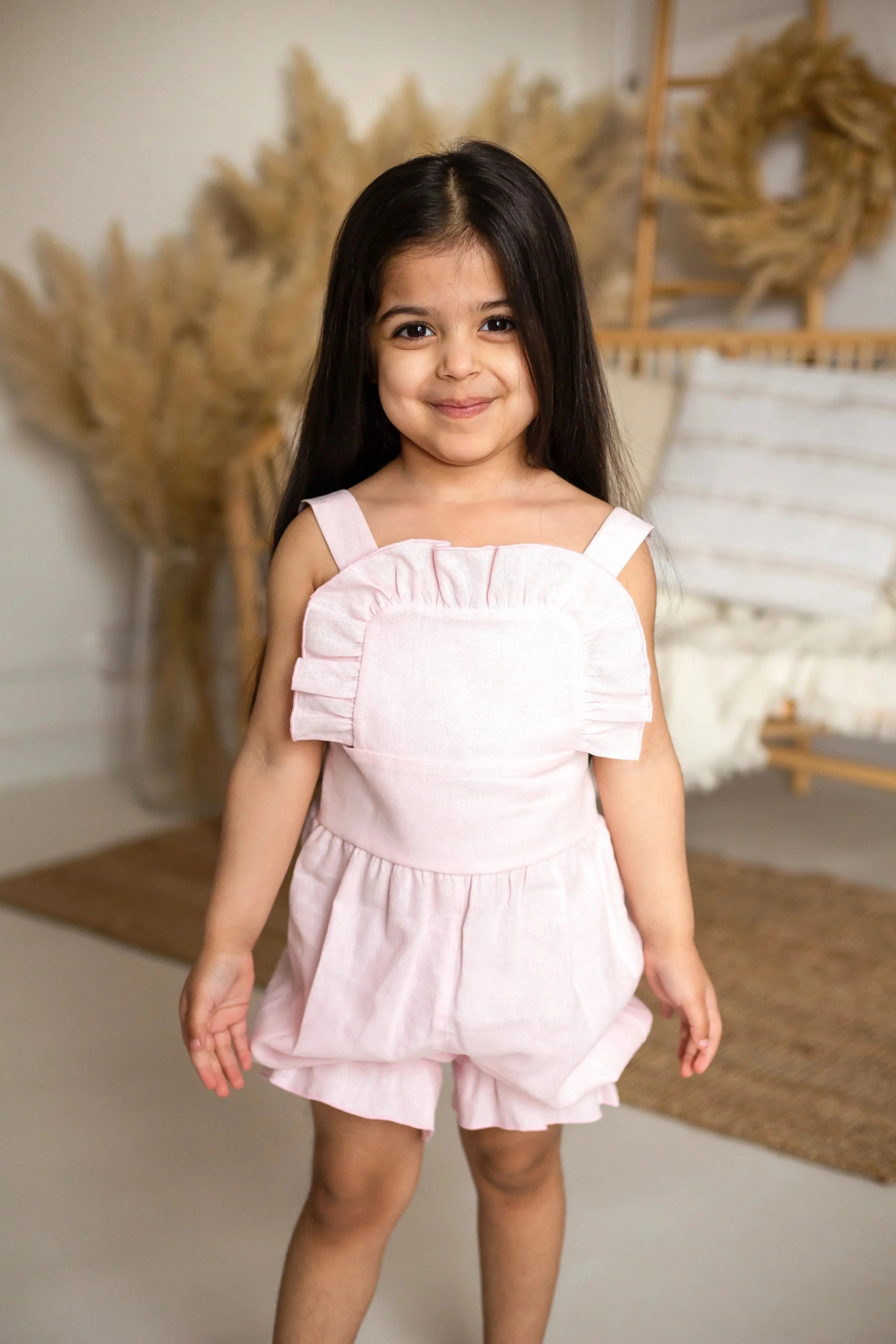 Smiling young girl in pink linen frilly jumpsuit in cozy home setting with neutral decor