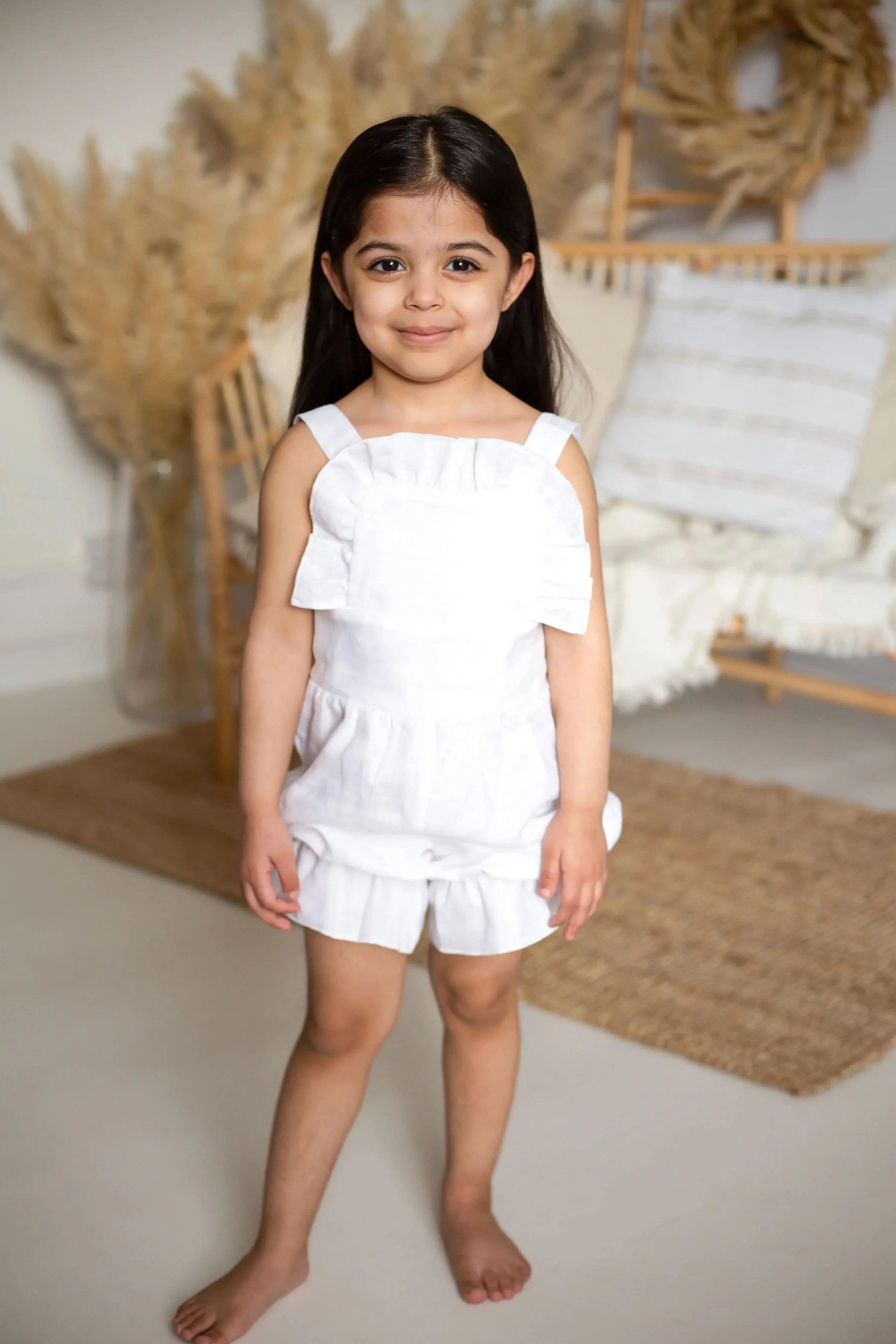 Smiling girl in an ivory linen frilly jumpsuit standing barefoot in a cozy, neutral-toned room