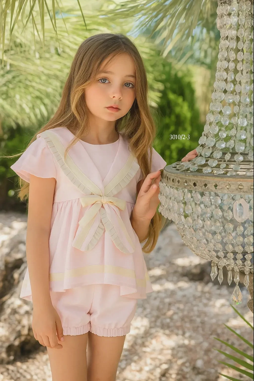 Young girl wearing a pink sailor short set with bow detail, standing outdoors by a crystal chandelier