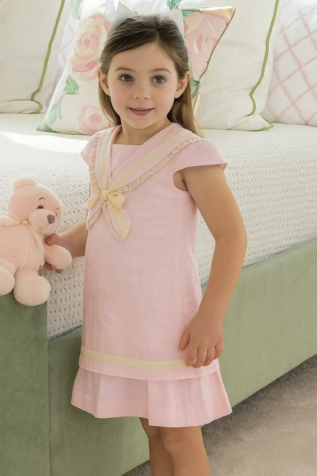Little girl wearing a pink linen sailor A-line dress holding a teddy bear in a cozy bedroom