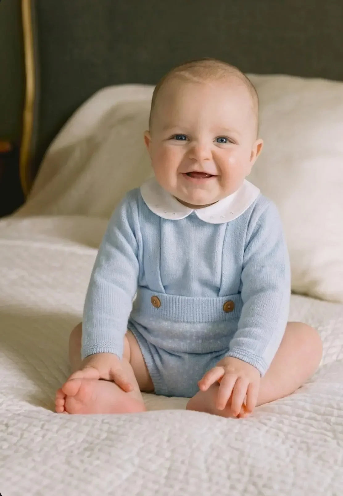 Smiling baby in blue knit outfit sitting on white quilted bed with gray headboard