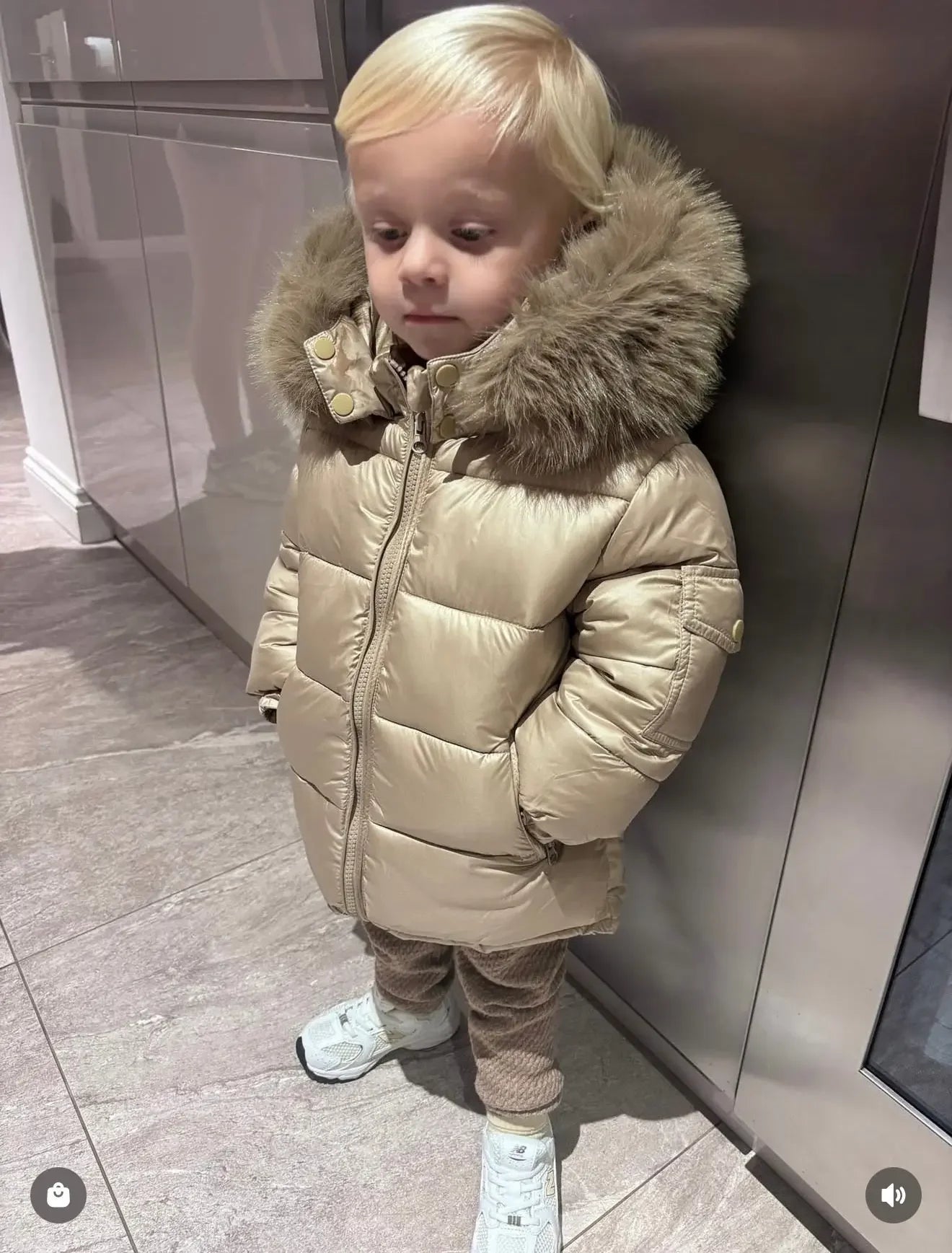 Toddler in a beige puffer jacket with fur hood, beige pants, and white sneakers standing indoors