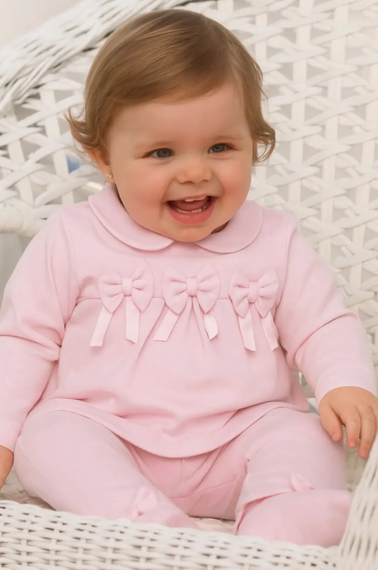 Smiling baby girl in pink outfit with bows sitting on white wicker chair