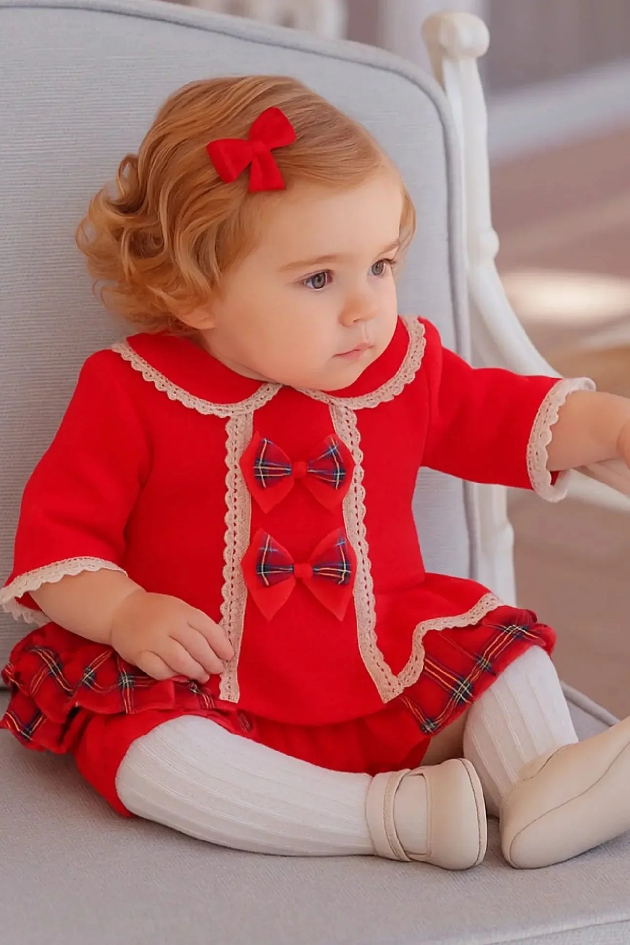 Toddler girl sitting on gray chair wearing red dress with plaid bows and white tights