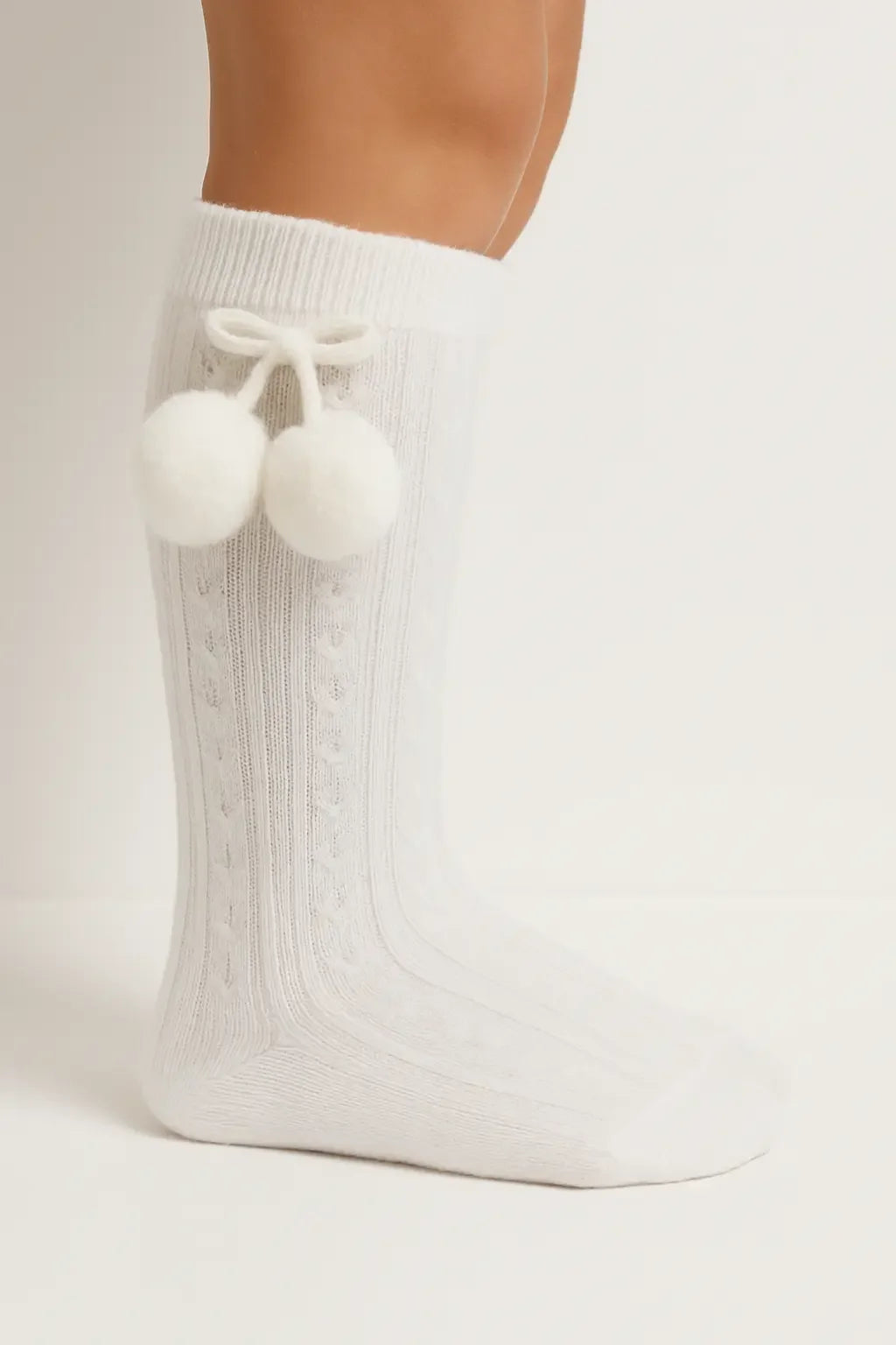 Close-up of leg wearing white cozy knit socks with pom-pom detail on neutral background