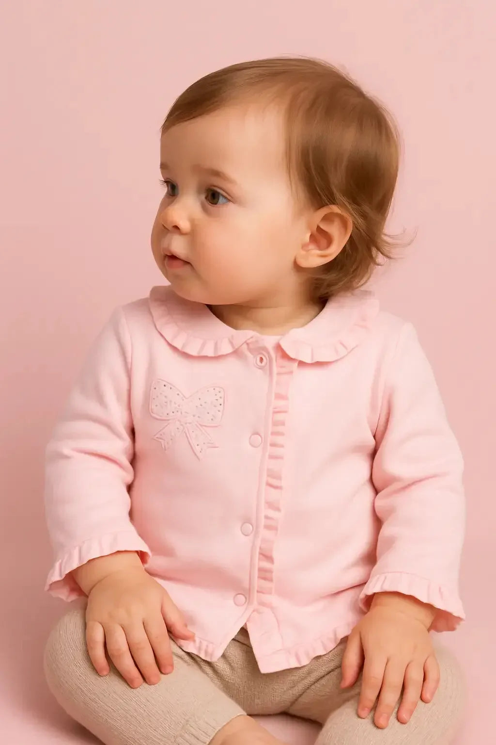 Cute baby girl in pink ruffled cardigan and beige pants sitting against pink background