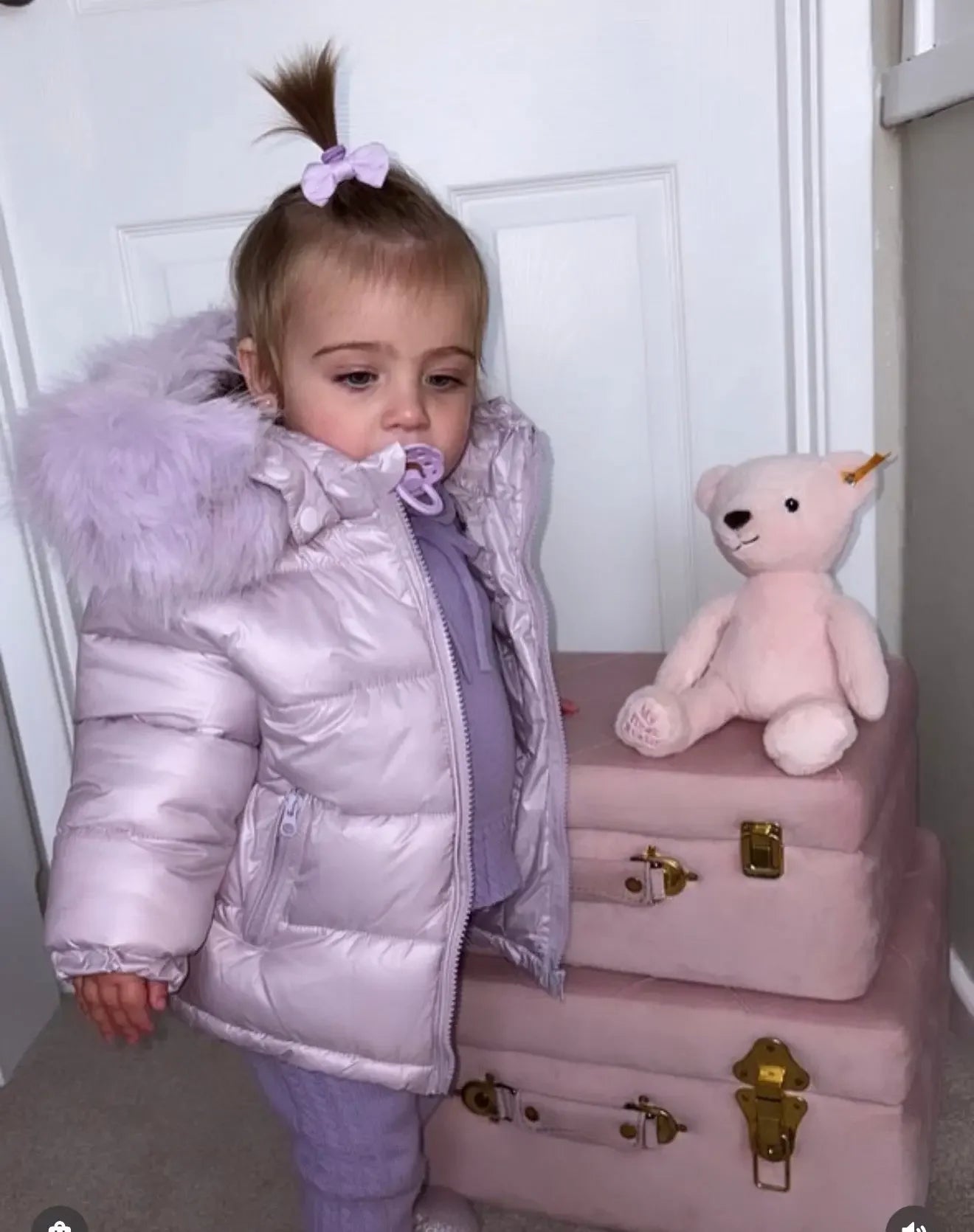 Toddler girl in light purple puffer jacket and matching outfit with pacifier, next to pink velvet suitcases and teddy bear
