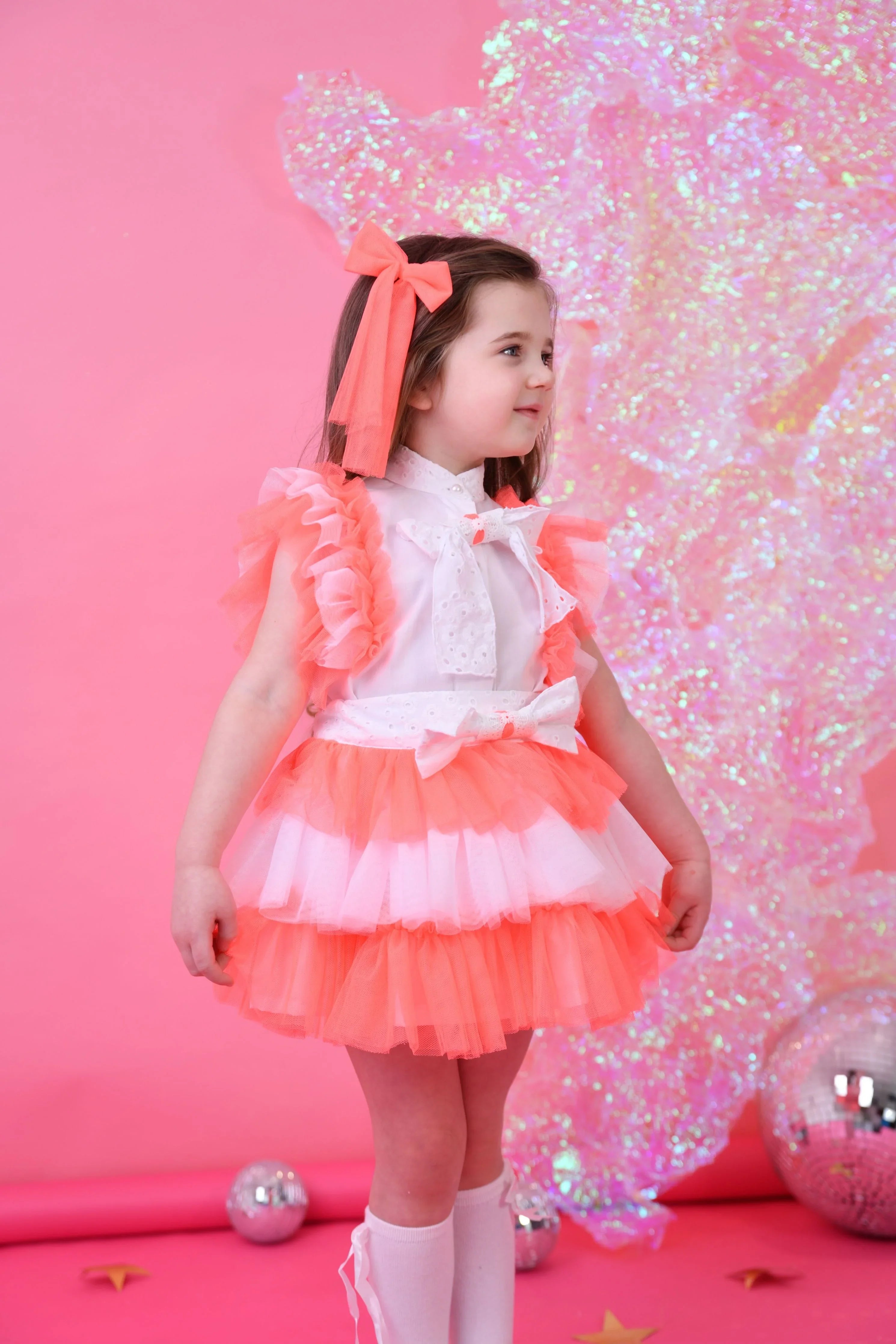 Young girl in neon coral and white tulle skirt set with ruffled sleeves and matching hair bow on pink backdrop