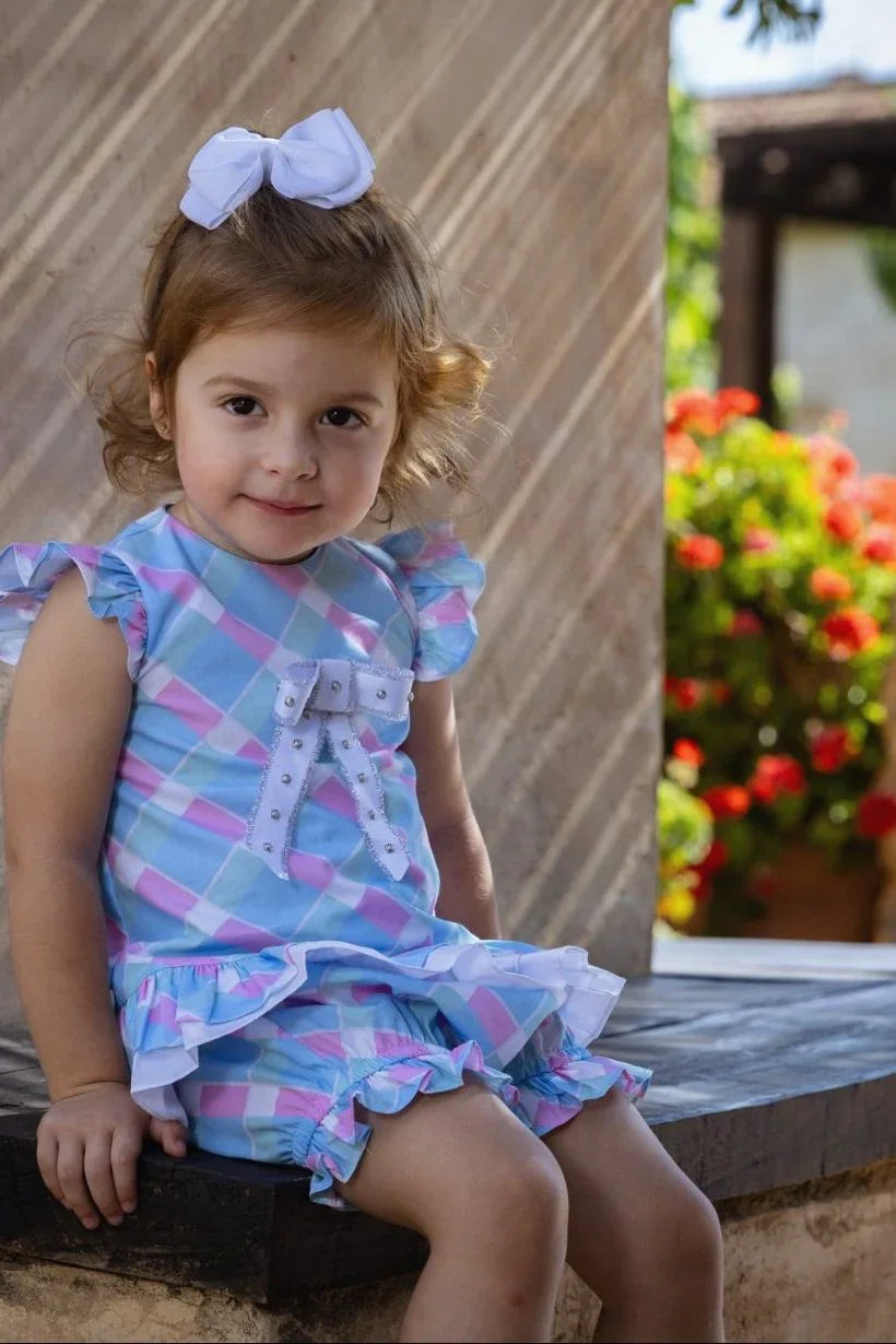 Toddler girl wearing pastel blue and pink plaid romper with ruffles and white bow sitting outdoors near flowers