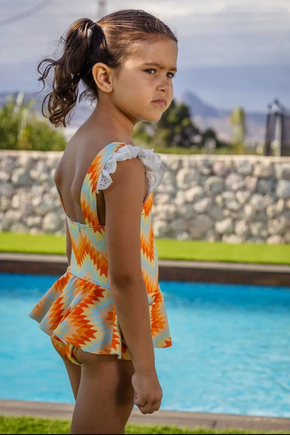 Young girl with ponytail wearing orange and blue zigzag swimsuit with white lace, standing by a pool