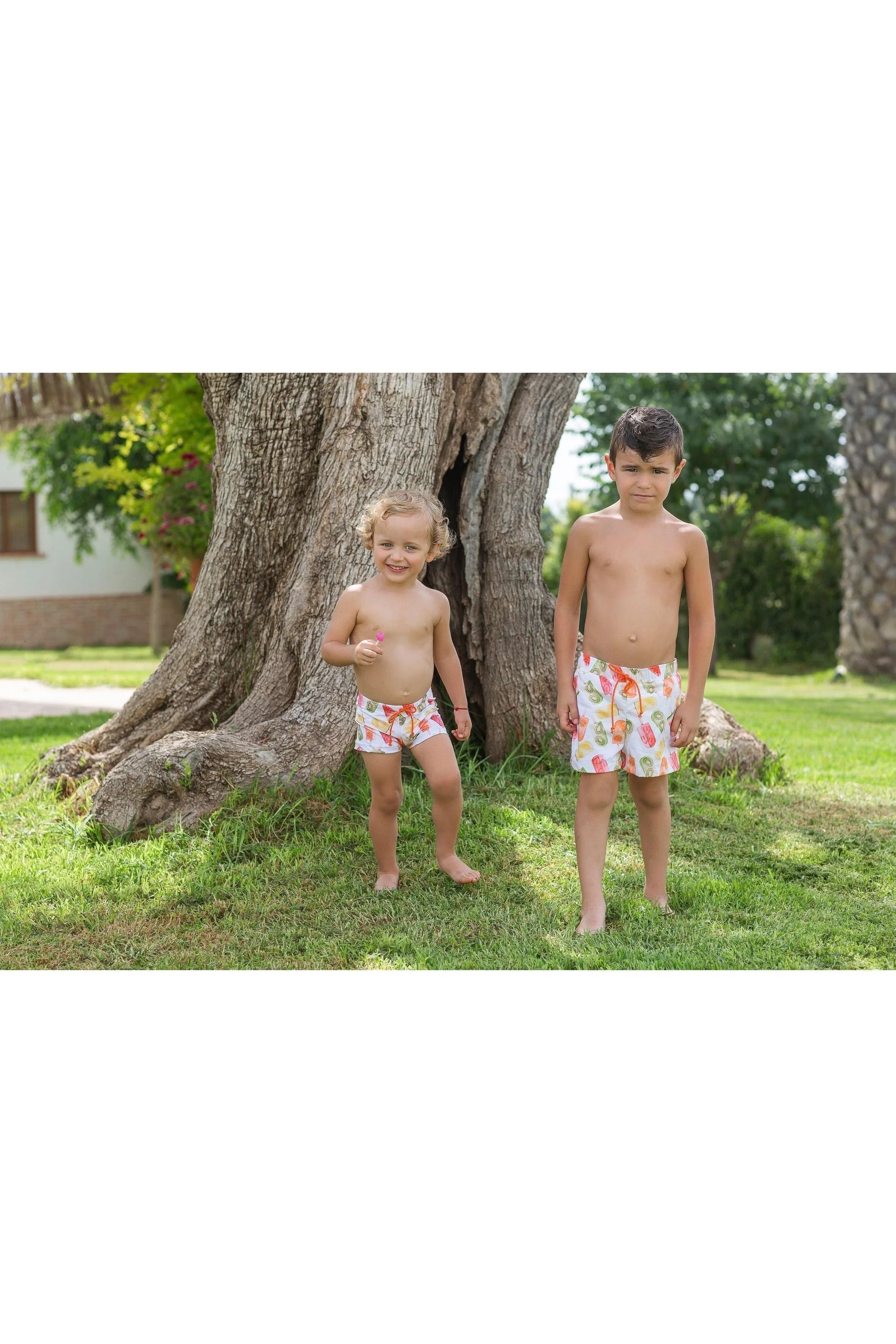 Two boys wearing colorful swim shorts with fruit print standing barefoot on grass near large tree