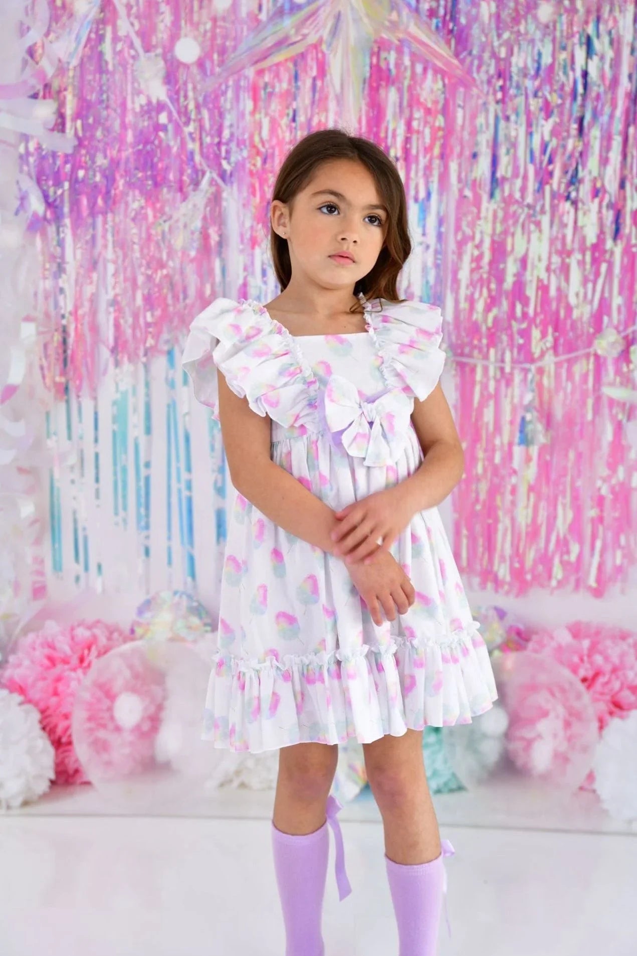 Young girl wearing a pastel frill dress and lavender socks against a pink and white party backdrop