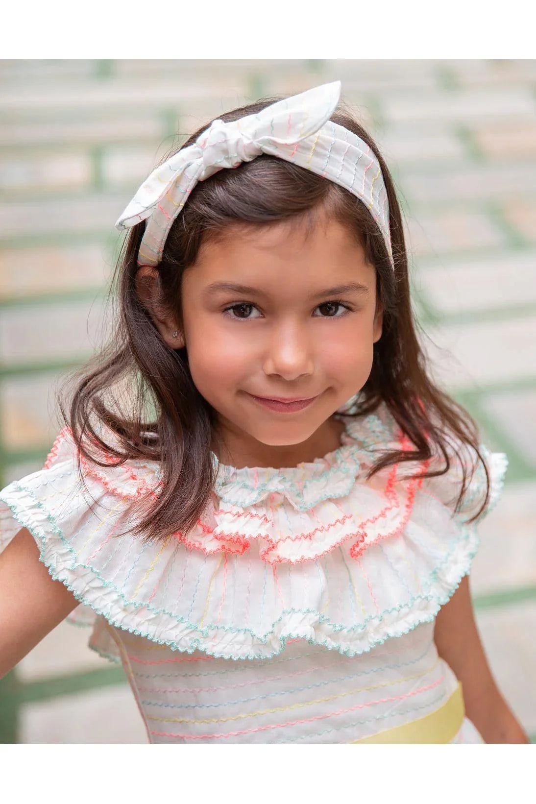 Smiling girl wearing white striped ruffled dress and matching bow headband in soft outdoor setting