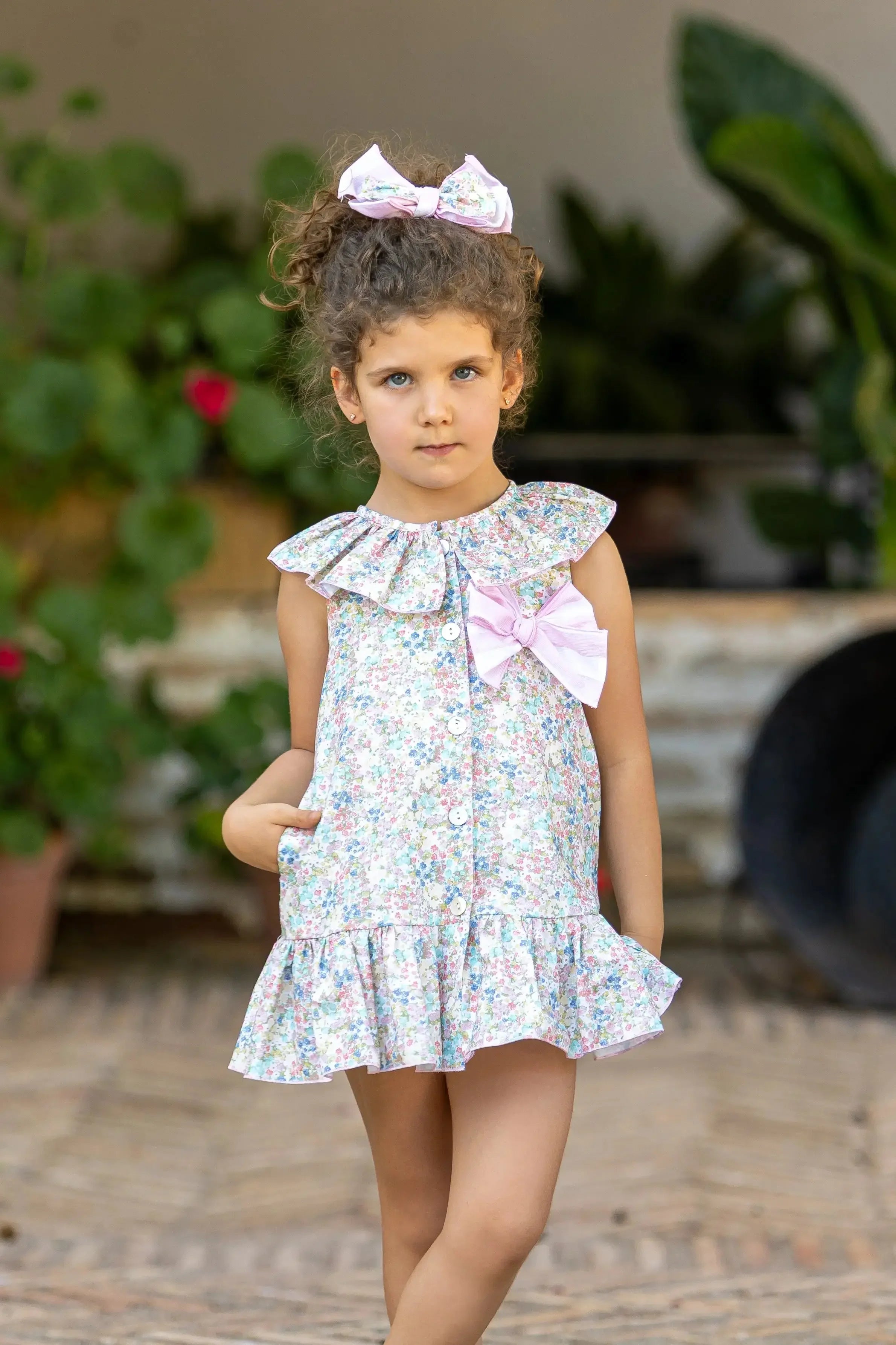 Young girl wearing pink floral dress with ruffled collar and bow, standing outdoors near plants
