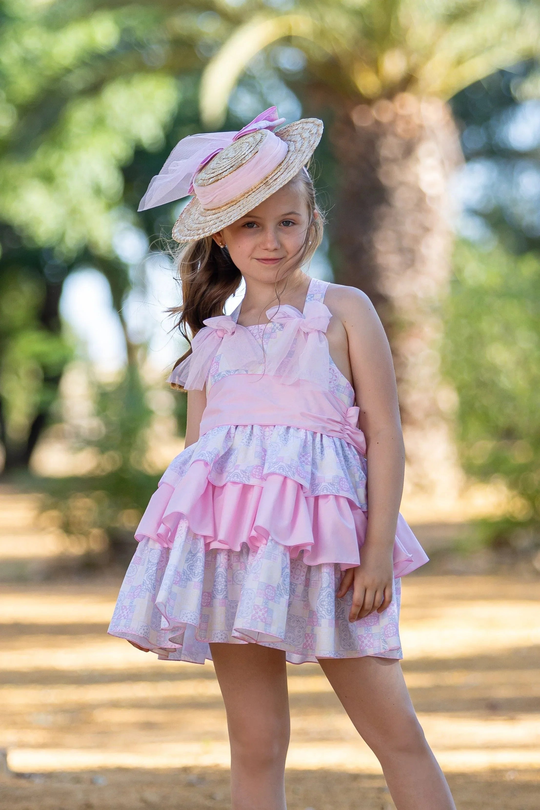Young girl wearing pink tiles dress and straw hat with pink ribbon outdoors near palm trees