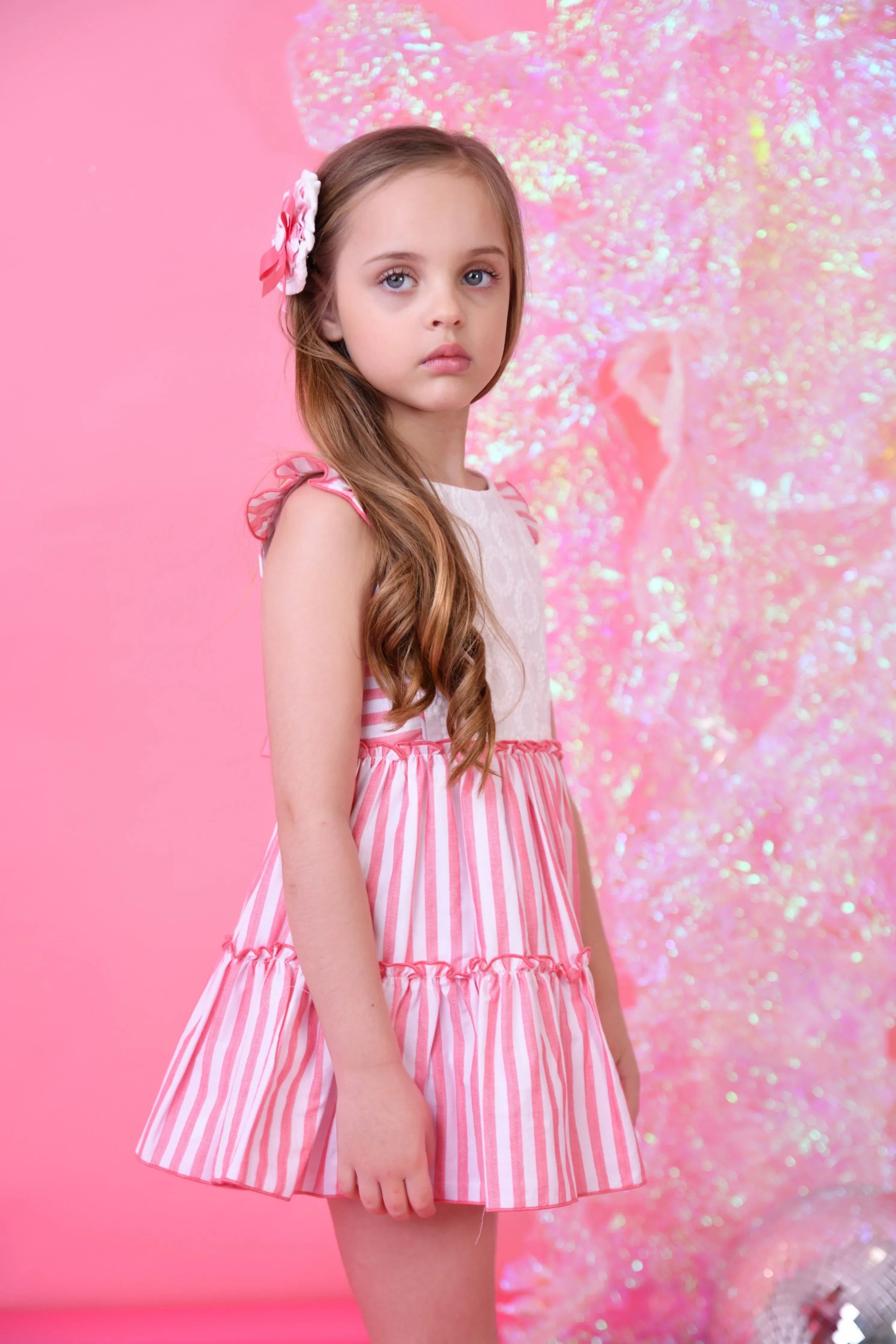Young girl in pink and white striped summer dress with flower hair clip against pink glittery background