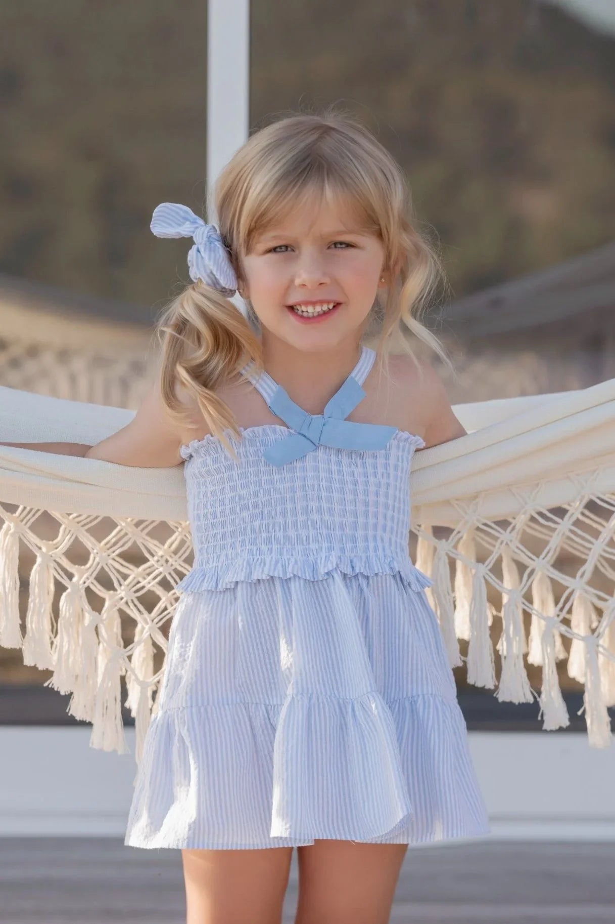 Little girl in blue and white striped sundress standing with arms on hammock outdoors