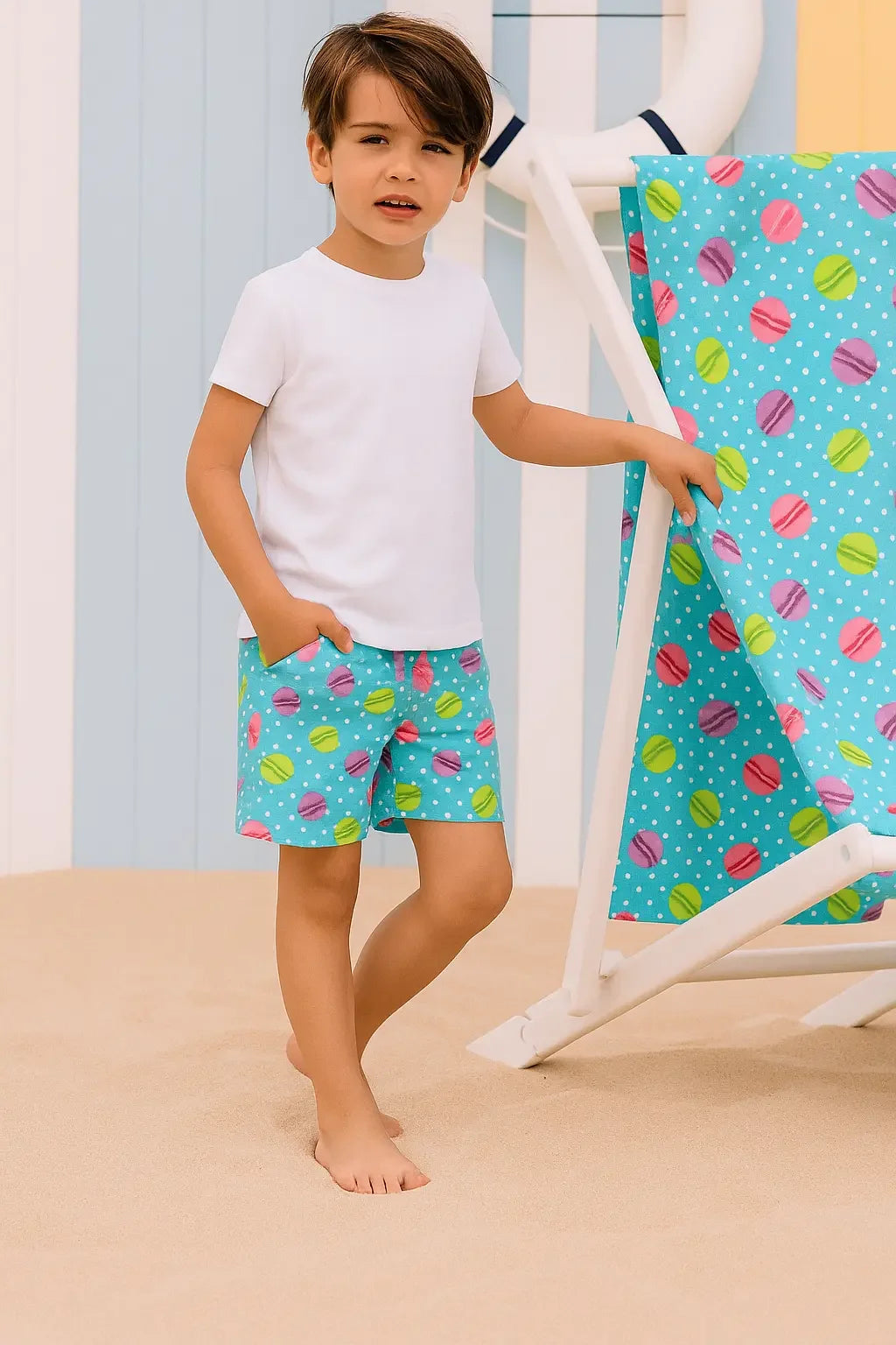 Young boy in white t-shirt and colorful polka dot swim shorts standing barefoot on sand next to matching beach chair