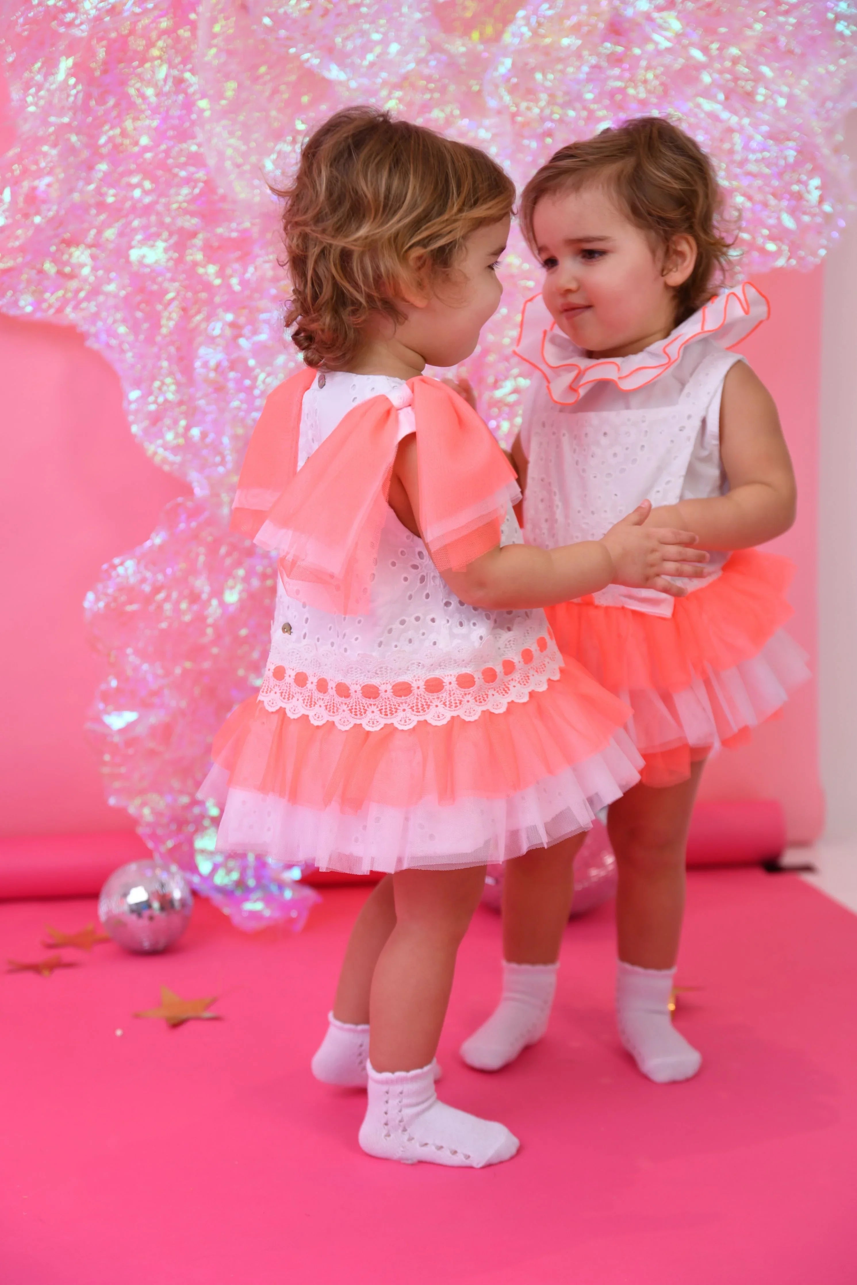Two toddler girls in pink and white tulle romper dresses on a pink background with iridescent decor