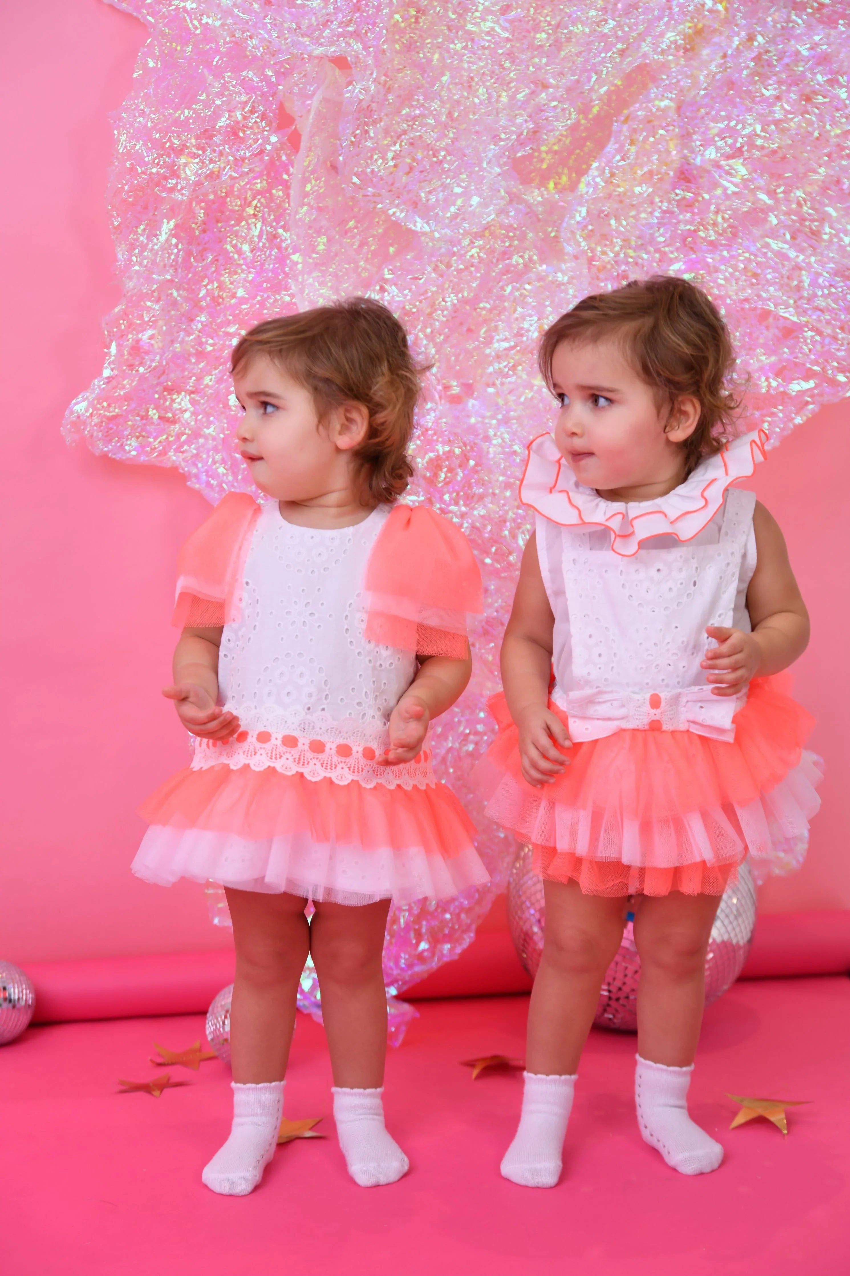 Two toddler girls in white and neon pink tulle romper dresses with white socks on a pink background
