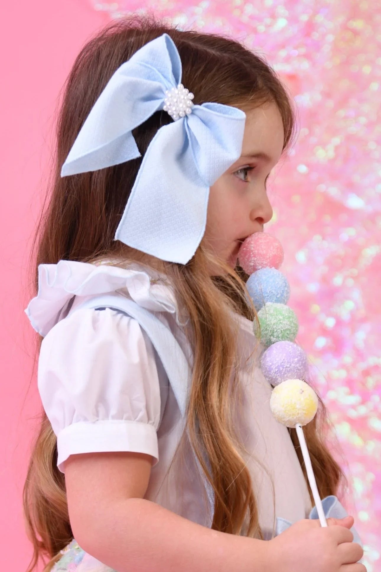 Young girl with long hair wearing a large blue bow hair clip and white ruffled blouse holding a pastel rainbow lollipop