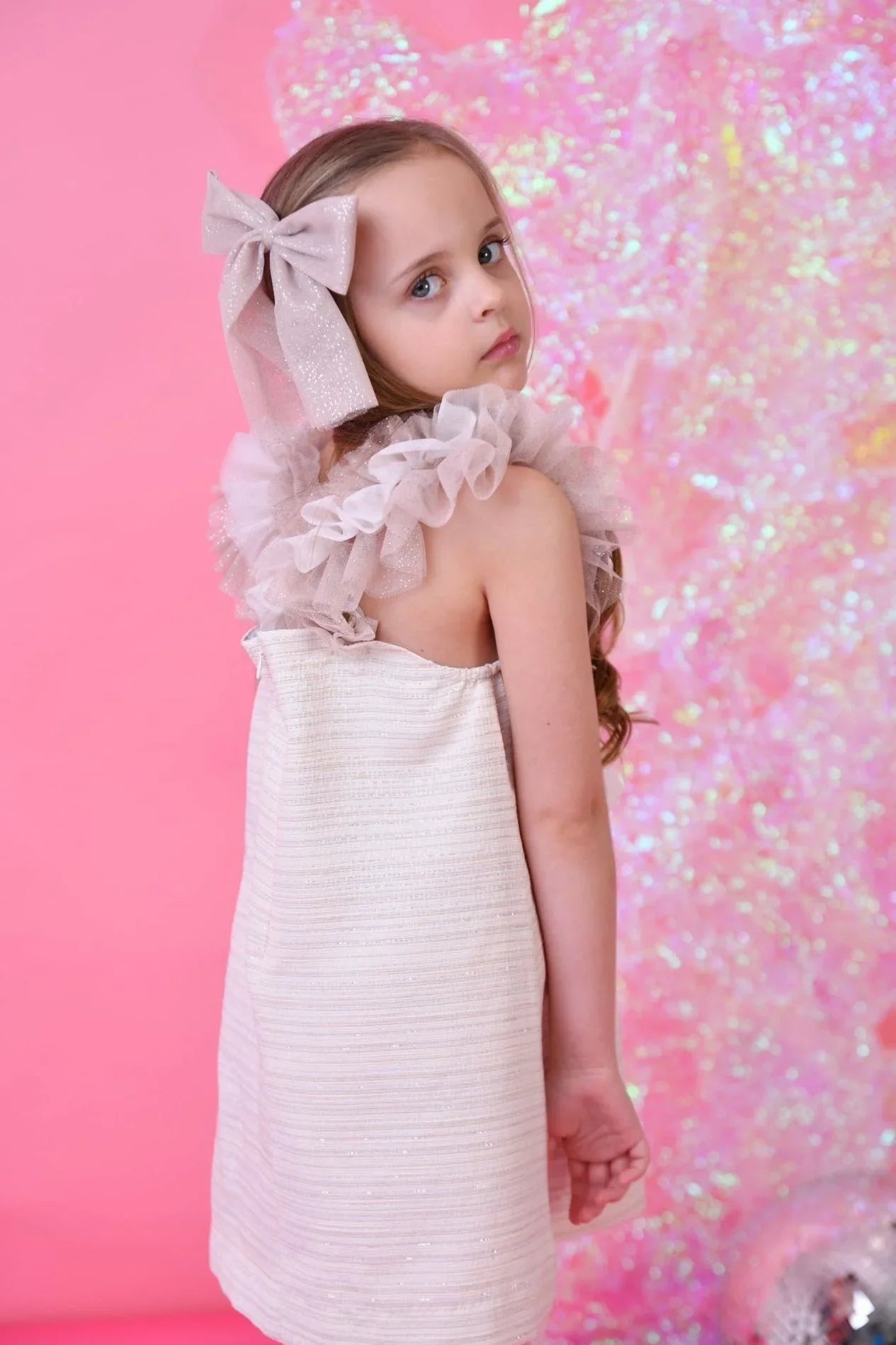 Young girl in a biscuit glitter tulle dress with a large bow in her hair against a pink background